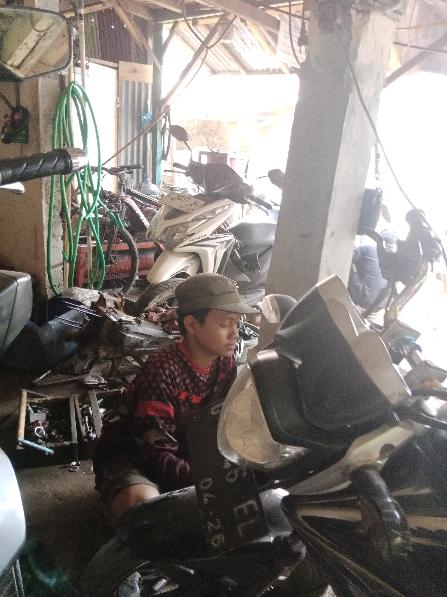 The image depicts a cluttered garage or workshop, likely located in Batang, Indonesia, given the context. In the foreground, a young person wearing a cap and a red and black patterned shirt is seated, appearing to be working on a motorcycle. Their hands are visible, suggesting they are actively engaged with the vehicle's engine parts, which are spread out on the floor. The motorcycle in the foreground is partially disassembled, with its engine exposed. Behind this person, another white Honda motorcycle is parked. Various tools and spare parts are scattered around the workshop, alongside gardening hoses and other miscellaneous items, indicating a busy and functional space. The overall environment is rustic, with a corrugated metal roof and wooden beams visible in the background. The lighting suggests it is daytime, with sunlight streaming in, creating some overexposed areas.