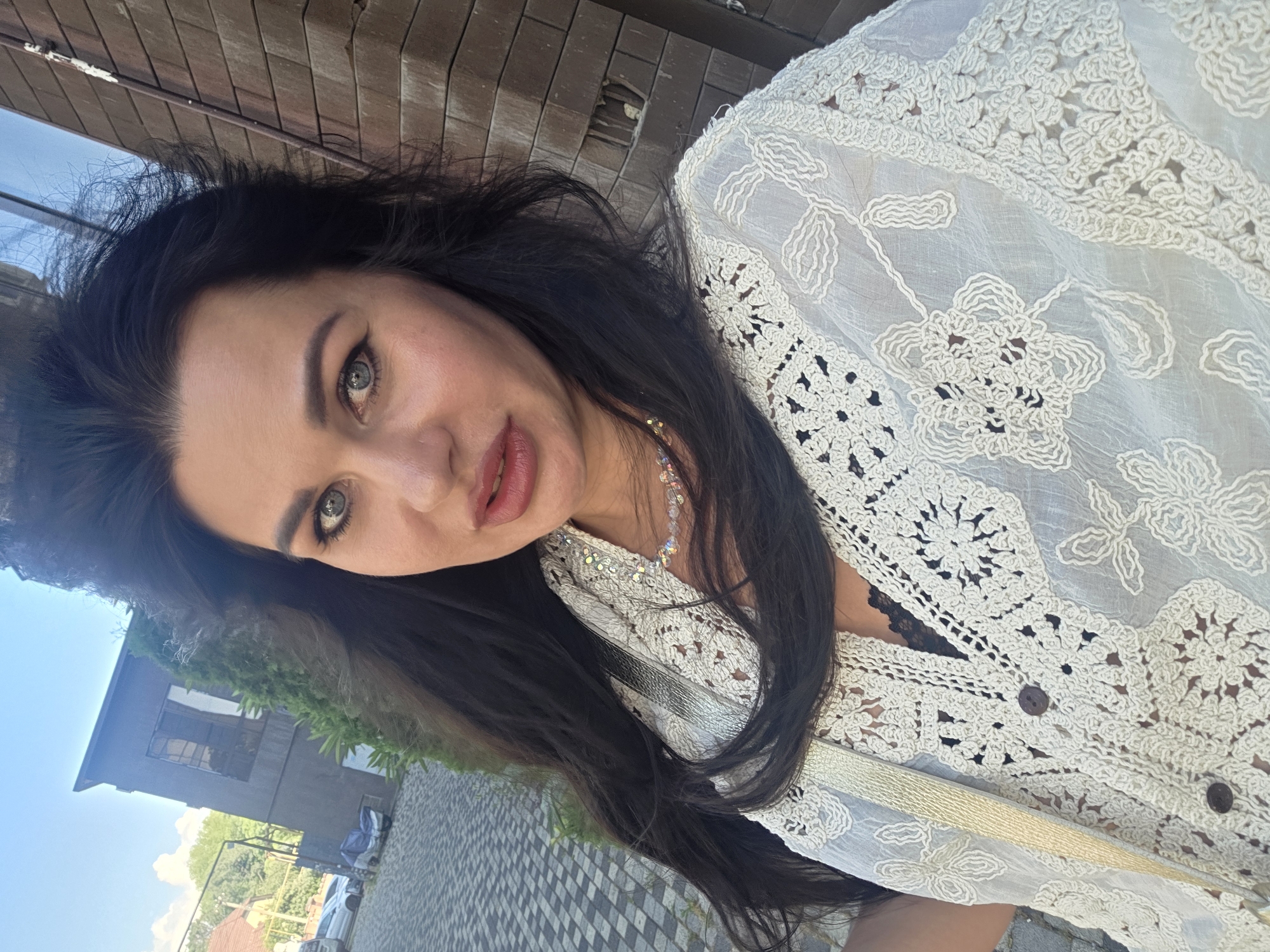 A woman with long, dark hair and blue eyes is pictured outdoors, looking at the camera. She is wearing a white, crochet-like garment over a black top, and a beaded necklace. She has on makeup, including lipstick and eyeliner. The background includes a brick building to the right and a paved area with some greenery and a building in the distance to the left. The setting appears to be an urban or suburban environment, possibly a courtyard or street. This scene is not specifically identifiable as being in Sochi, Russia based on visual cues alone.
