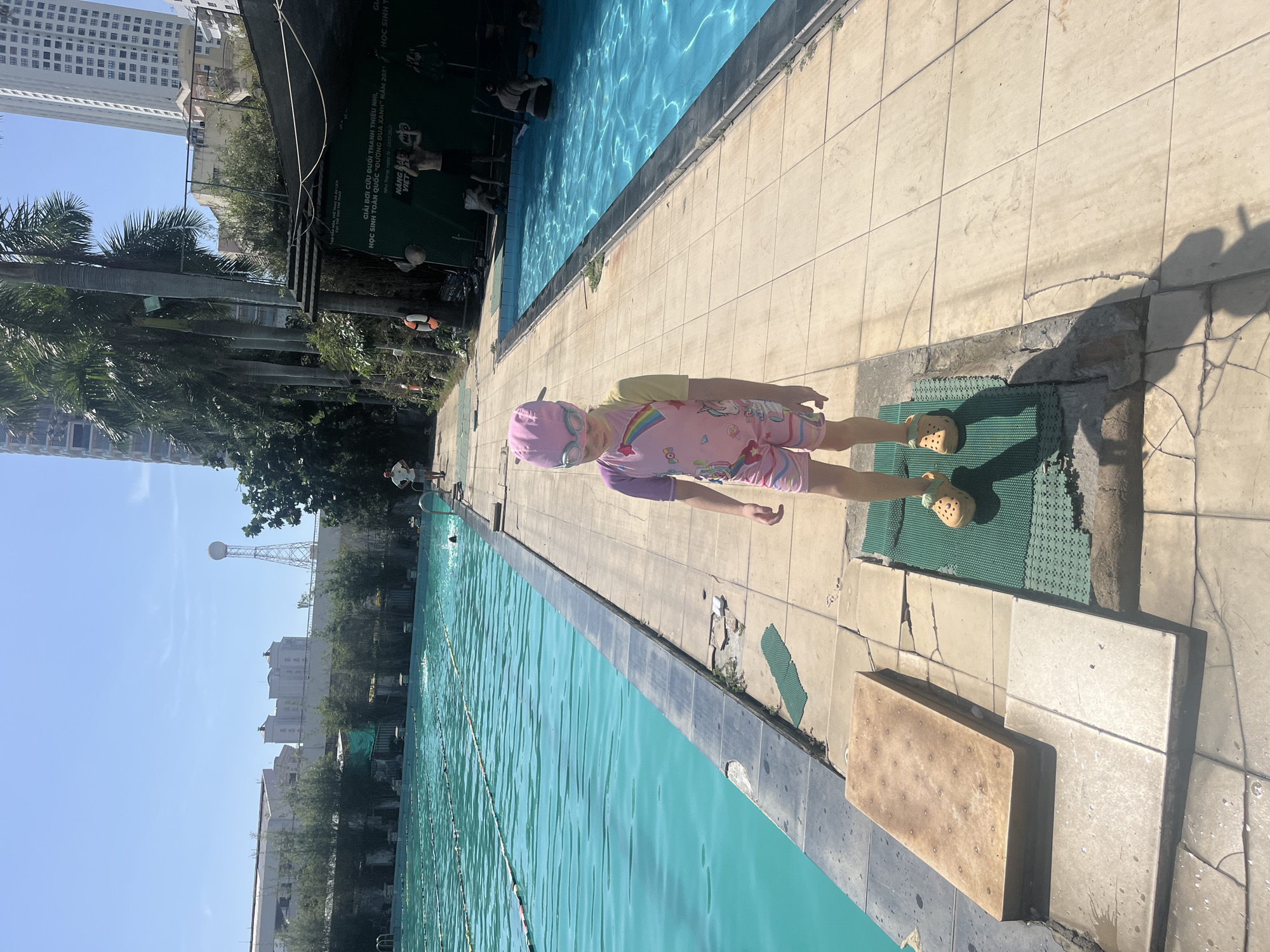 This image depicts an outdoor swimming pool complex located in Nha Trang, Vietnam, as evidenced by text on a green banner in the background. The scene is illuminated by bright sunlight, casting distinct shadows.
In the foreground, a young child stands centrally on a green textured mat. The child wears a pink and yellow full-body swimsuit with a rainbow and unicorn design, a pink swimming cap, light blue swimming goggles, and tan Crocs footwear with a smiling face emblem. The child is oriented towards the camera.
The complex features two swimming pools filled with turquoise water. A multi-lane pool extends across the left side of the frame, with lane ropes visible, and at least two individuals swimming within it. To the right, a smaller pool contains at least two individuals, one of whom is partially submerged and facing away.
The pools are bordered by a light-colored tiled concrete deck, some sections exhibiting cracks or repairs. Another green textured mat is partially visible to the child's right. A rectangular, flat, brown object rests on the deck near the edge of the larger pool.
Several other individuals are present on the deck. In the mid-ground right, a shirtless man wearing dark shorts stands near the smaller pool with his arms raised. Further back on the deck, another man in a white t-shirt and light shorts is visible. Another man in a white t-shirt and dark shorts stands on the deck in the background, to the left of the main subject. An orange lifebuoy is mounted on a pole near the right pool.
The background reveals an urban environment with various buildings, including tall modern high-rises and lower structures. Palm trees are visible along parts of the complex. A distinctive water tower with a spherical top on a lattice structure is present in the far background on the left. A large green banner with Vietnamese text, indicating an event held in Nha Trang from July 19-22, 2023, is positioned on the right side.