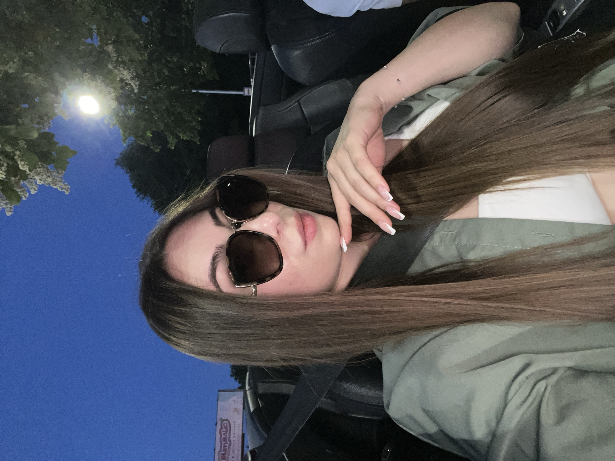 A young woman with long brown hair and sunglasses is in the passenger seat of a convertible car at night. She is wearing a green jacket over a white top. Her left hand is resting on her chin, showing off her French manicured nails. The car's interior is visible, with black leather seats and a seatbelt. In the background, trees and a street light can be seen, suggesting an outdoor urban setting. A billboard is partially visible on the left. The sky is dark blue. The image appears to have been taken in the city of Plovdiv, Bulgaria, possibly near a commercial area given the billboard.