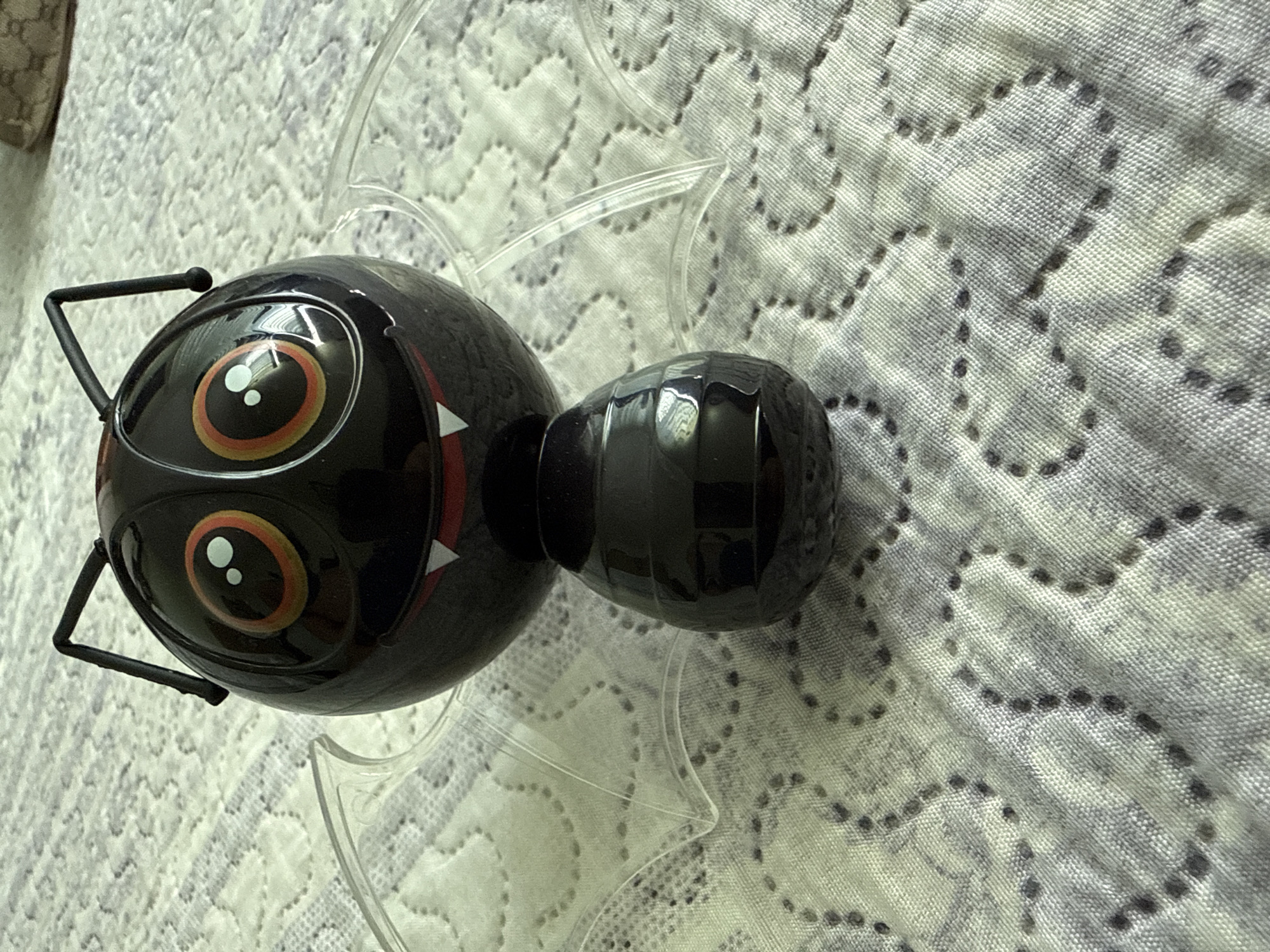A black, bug-shaped toy with large, cartoonish eyes and clear wings sits on a white quilted blanket. The toy has two black antennae extending from its head. Its mouth is open, revealing small white fangs. The blanket has a subtle pattern of grey dotted lines forming a wave-like design. The scene is indoors. The image does not provide any information about the city of Ekzarh Antimovo, Bulgaria.