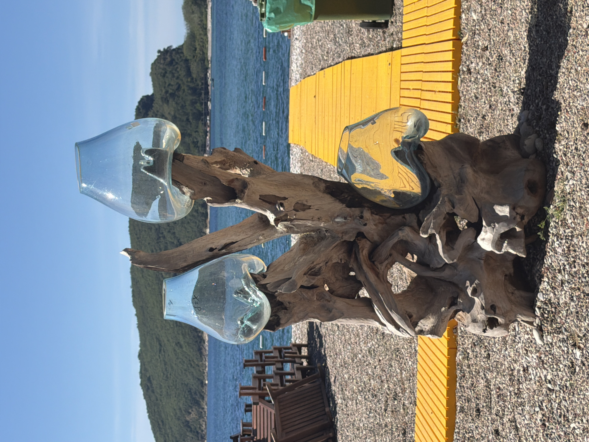 Here is a detailed description of the image:
**Scene:** An outdoor scene with a decorative driftwood sculpture on a beach.
**Location:** Likely Budva, Montenegro, based on the coastal landscape.
**Objects:**
* A large piece of driftwood, shaped into an artistic sculpture.
* Three clear, blown glass vases or containers mounted on the driftwood.
* A yellow boardwalk-style pathway is visible to the right of the sculpture and near the top of the frame.
* A pebble beach covers the ground.
* In the background, the sea is visible, along with land covered in green vegetation.
**Environment:** A sunny day.
*The image is rotated.