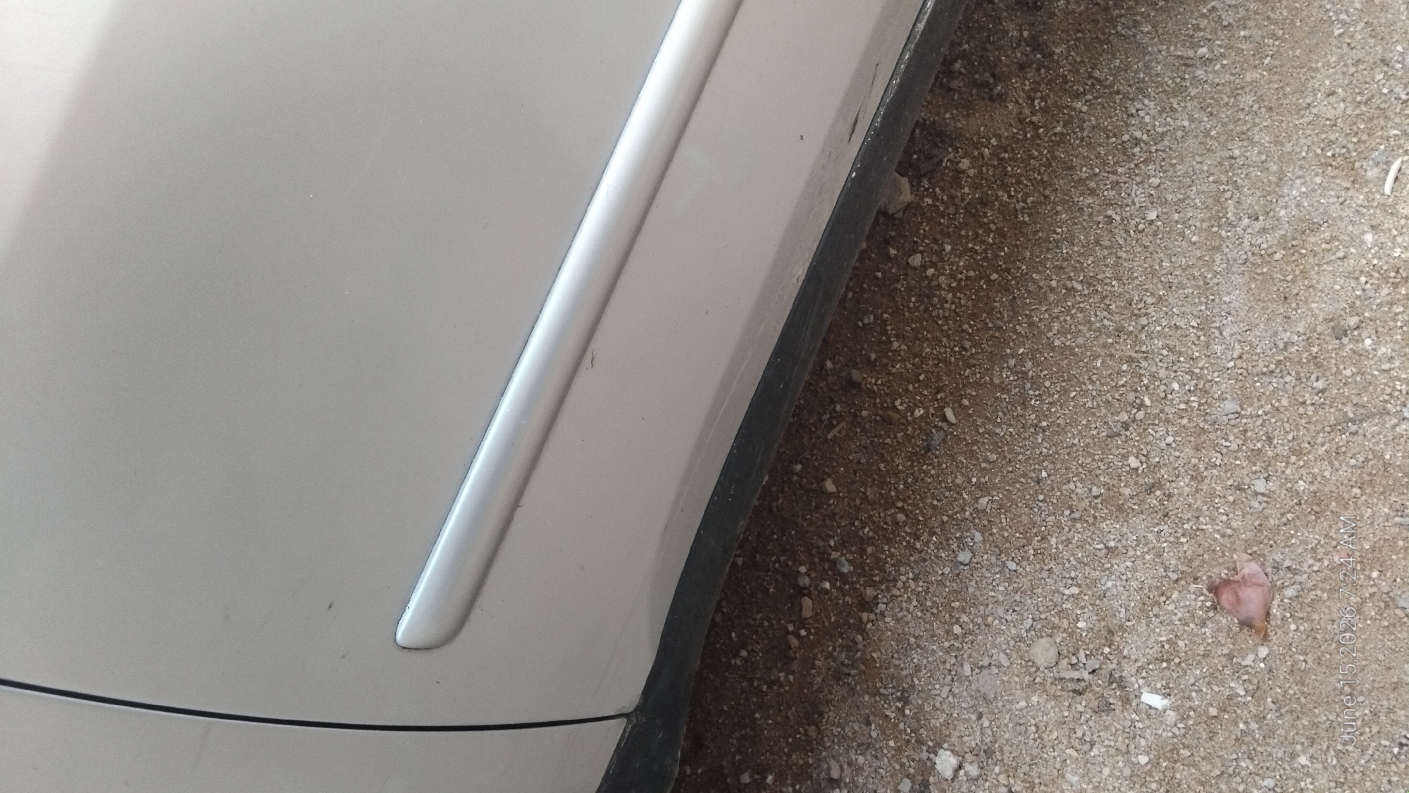 The image shows a close-up view of the lower portion of a silver car door and the ground beneath it. The car has a silver trim strip running horizontally along the door. The ground is composed of dirt and small pebbles, with a dark black plastic trim piece visible along the bottom edge of the car. A small, reddish-brown leaf or piece of debris rests on the ground near the bottom left of the image. The background is mostly out of focus, suggesting the photo was taken outdoors. The timestamp in the bottom left corner indicates "June 15, 2026, 7:24 AM." There are no people or actions visible in the image. The scene appears to be a typical street or parking area, and there is no specific indication of it being located in Howayi, Nigeria, other than the information provided in the prompt.