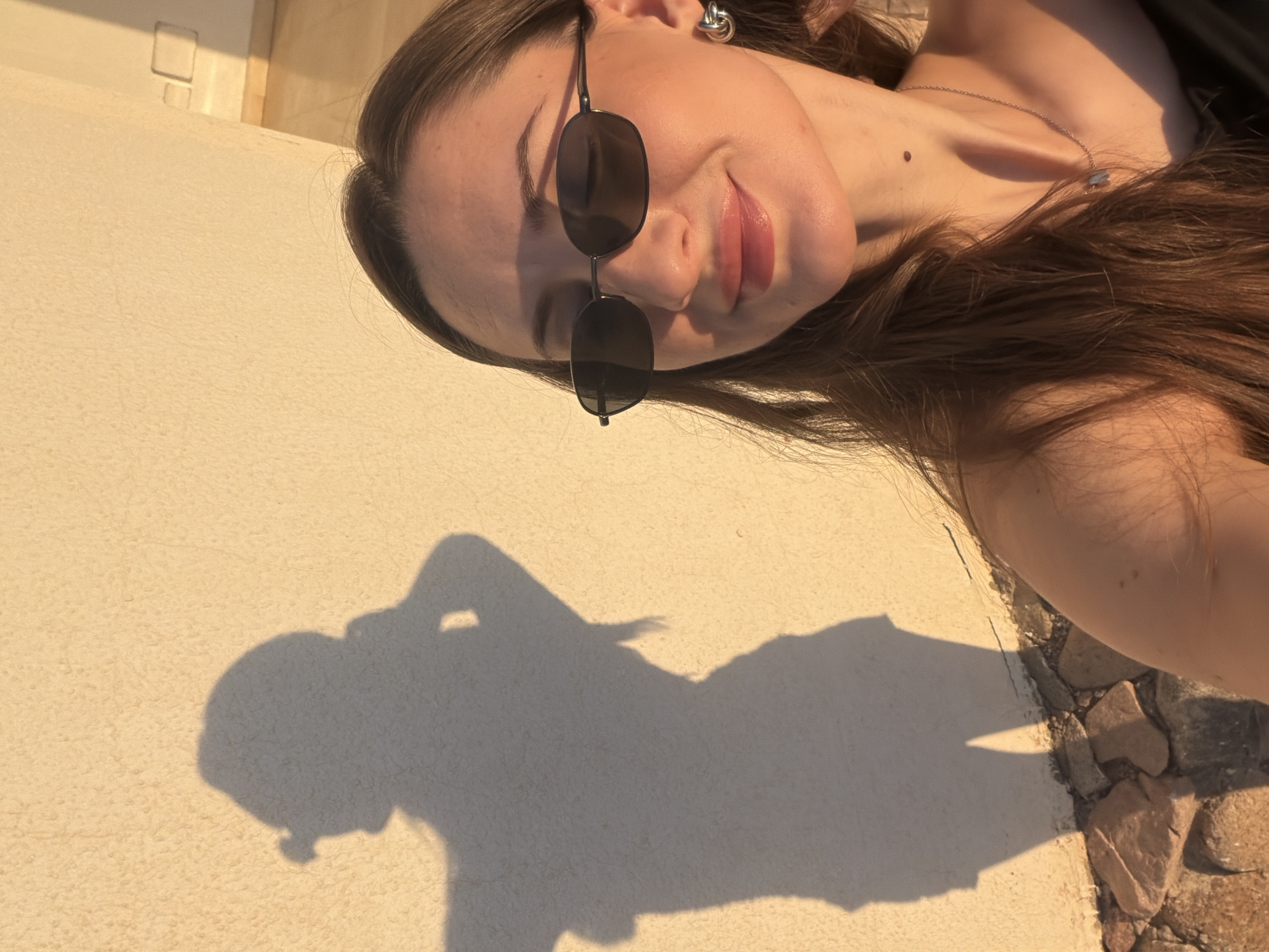 An unidentifiable young woman, facing the camera and wearing sunglasses and a dark top, is positioned in the foreground, with her shadow cast on a textured, light-colored wall behind her. The woman's right shoulder and chest are visible, along with her long, brown hair, which is falling over her shoulder. She is also wearing a silver necklace with a pendant and a silver hoop earring in her right ear. The shadow on the wall depicts a full-body silhouette of a person in a similar pose, suggesting the original image was taken at a distance to capture both the subject and her shadow. The background wall is illuminated by warm, golden sunlight, indicating it is likely late afternoon or early morning. The lower portion of the wall meets a foundation made of rough, natural stones. The overall setting appears to be an outdoor location, possibly a terrace or courtyard, given the presence of the wall and the sunlight. The image does not provide enough context to definitively place it within Sharm El Sheikh, Egypt, though such an environment is plausible for the region.