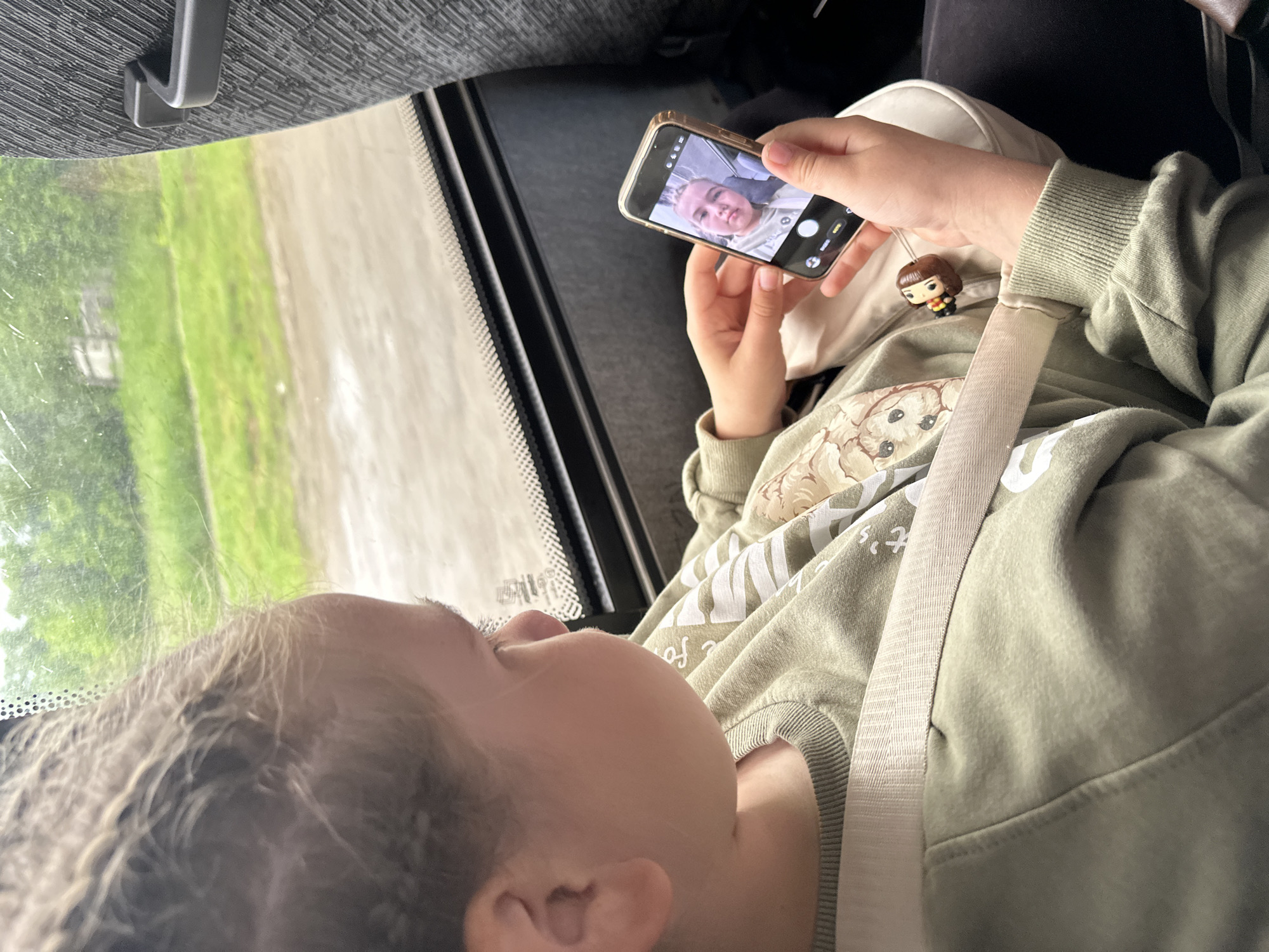 A young child with light-colored hair, secured by a cream-colored seatbelt, is seated within a vehicle. The child wears an olive-green long-sleeved sweatshirt adorned with white graphics, some of which appear to be Japanese characters. In both hands, the child holds a gold-framed smartphone, its screen displaying the front-facing camera view, showing a selfie of the child's own face within the frame, indicating active use of the camera function in "PHOTO" mode. A small, dark-haired character charm, resembling a Funko Pop figure, is attached to a light-colored pouch near the child's right wrist. Through the vehicle's window, a blurred exterior landscape is visible, featuring green foliage, grassy areas, and a dirt or unpaved road. The vehicle interior shows dark window frames and a textured grey seat. The specific location of Кварса, Russia, cannot be determined from the visual information present in the image.
