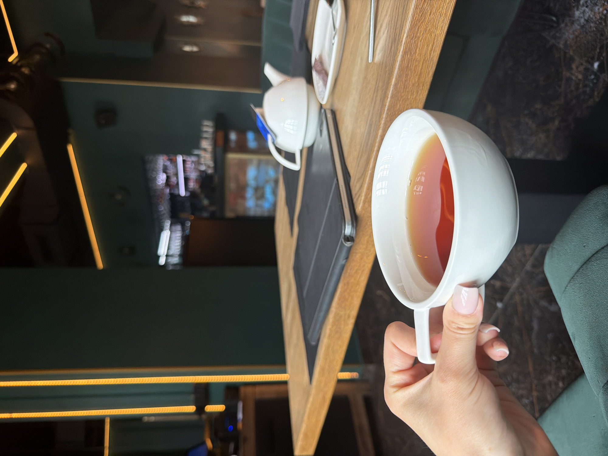 A close-up, portrait-orientation shot captures a hand holding a cup of tea in what appears to be a restaurant or cafe setting.
The main focus is on a white teacup filled with a dark liquid, presumably tea. The cup is held by a hand with manicured nails, suggesting the subject is likely a female.
In the background, there's a wooden table with a teapot and a saucer. A menu or bill presenter is also visible. The background is a mix of blurred interior details, with some light fixtures and a hint of a television screen. The setting appears to be a cafe or restaurant with a modern design.