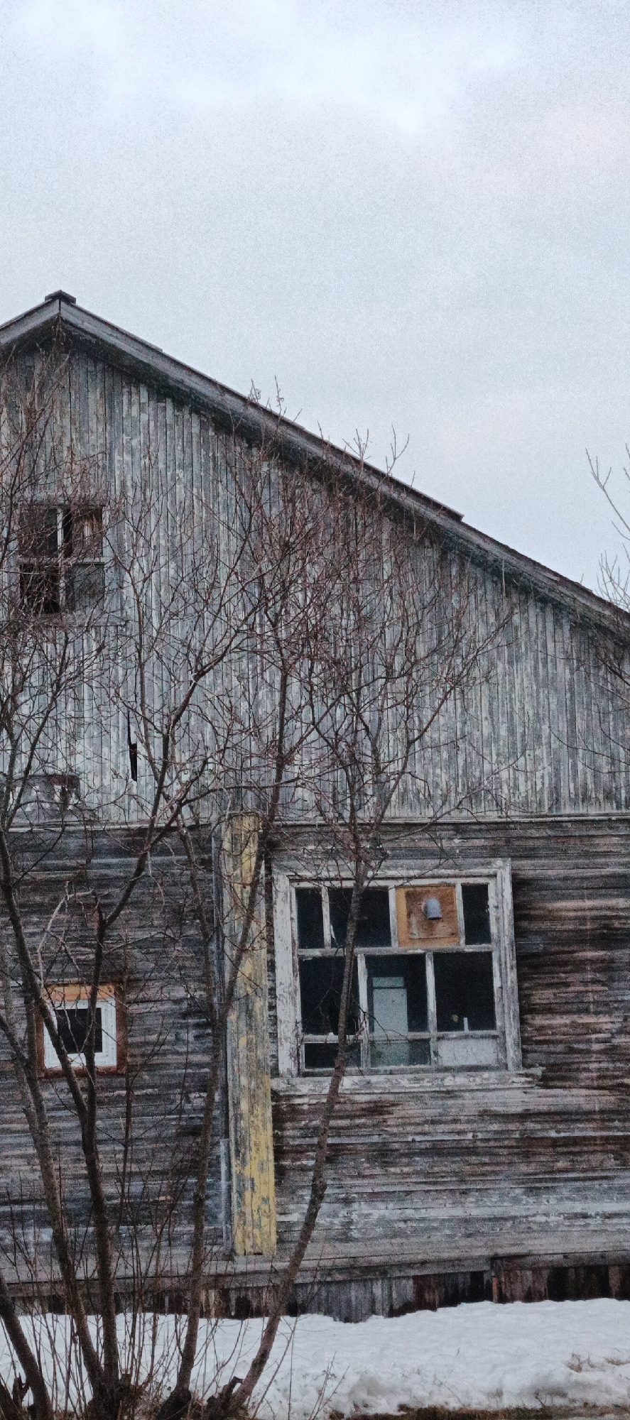 This image depicts a two-story, heavily weathered wooden building located in Nar'yan-Mar, Russia. The structure features a dark gable roof and vertical gray plank siding on its upper level. A small, dark, rectangular window is present on the upper left, alongside a narrow vertical opening. The lower level utilizes horizontal gray plank siding, with a central vertical section exhibiting peeling yellow paint. A large, multi-pane window frame on the lower right shows multiple broken or absent glass panes; its central upper section is boarded with a light brown material, fitted with a small, bell-shaped fixture. A smaller, rectangular window with a light frame is visible on the lower left. Dormant, bare-branched trees and shrubs partially obscure the building in the foreground. The ground is covered by patchy, discolored snow. The sky is uniformly overcast and light gray. No human or animal subjects are present.