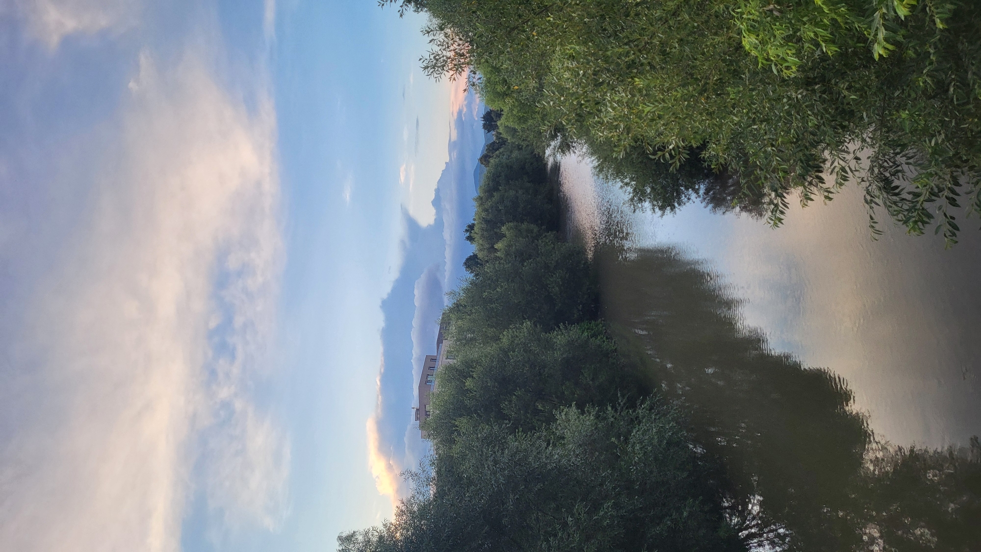 The image depicts a wide, muddy river flowing through a city, lined with dense, dark green trees on both banks. The river reflects the sky and the surrounding foliage. In the background, a multi-story building with a light-colored facade is visible above the trees, and in the far distance, mountains are silhouetted against a hazy sky. The sky itself is a mixture of light blue and pastel-colored clouds, suggesting either early morning or late evening light. The overall scene evokes a sense of peaceful, natural beauty within an urban setting. This scene is likely located along the Sekčov River in Prešov, Slovakia, given the presence of urban development adjacent to the river and the typical landscape of the region.