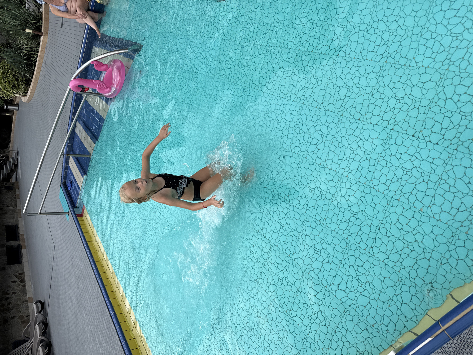 This is a high-angle, horizontal, outdoor, full shot of a young girl playing in a swimming pool. The girl, with blonde hair, is wearing a black and white patterned swimsuit. She is in the middle of the pool, splashing water with her legs and arms as if jumping or playing. Her left arm is extended upwards, and her right arm is bent. She has a red bracelet on her right wrist. Her expression is not clearly visible.
The pool is filled with bright turquoise water, and the bottom is tiled with a mosaic pattern of irregular, greyish-blue shapes outlined in black. The pool has a yellow and blue tiled edge. To the left of the pool, there is a grey concrete deck with a metal railing. On the deck, partially submerged in the water, is a bright pink flamingo-shaped inflatable toy. In the upper left corner, out of focus, a person wearing a striped swimsuit is sitting on the edge of the pool. The scene appears to be set during the daytime, likely on a sunny day, given the bright lighting. The overall mood seems to be one of fun and recreation. There is no visible text in the image. The location context provided is Массандровский поселковый совет, Ukraine, suggesting a possible location for the swimming pool.