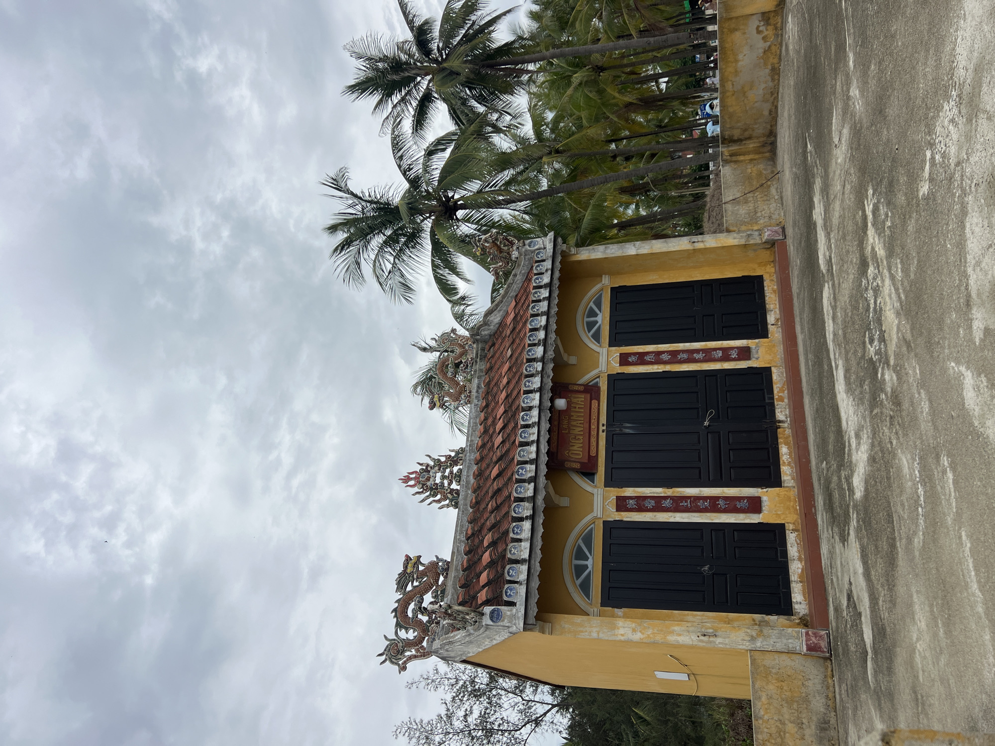 The image depicts a yellow building with a tiled roof adorned with dragon figurines, located in Hoi An, Vietnam. The building has three large black doors and two arched windows above them. Red banners with Chinese characters are displayed on the building's facade. Several palm trees are visible behind the building, suggesting a tropical or subtropical environment. The sky is overcast, with scattered clouds. The ground in front of the building is paved with concrete. In the background, beyond the palm trees, some people and a vehicle can be seen. The overall scene suggests a cultural or historical site. The building's design, particularly the dragon motifs and the architectural style, are characteristic of traditional Vietnamese temples or pagodas.