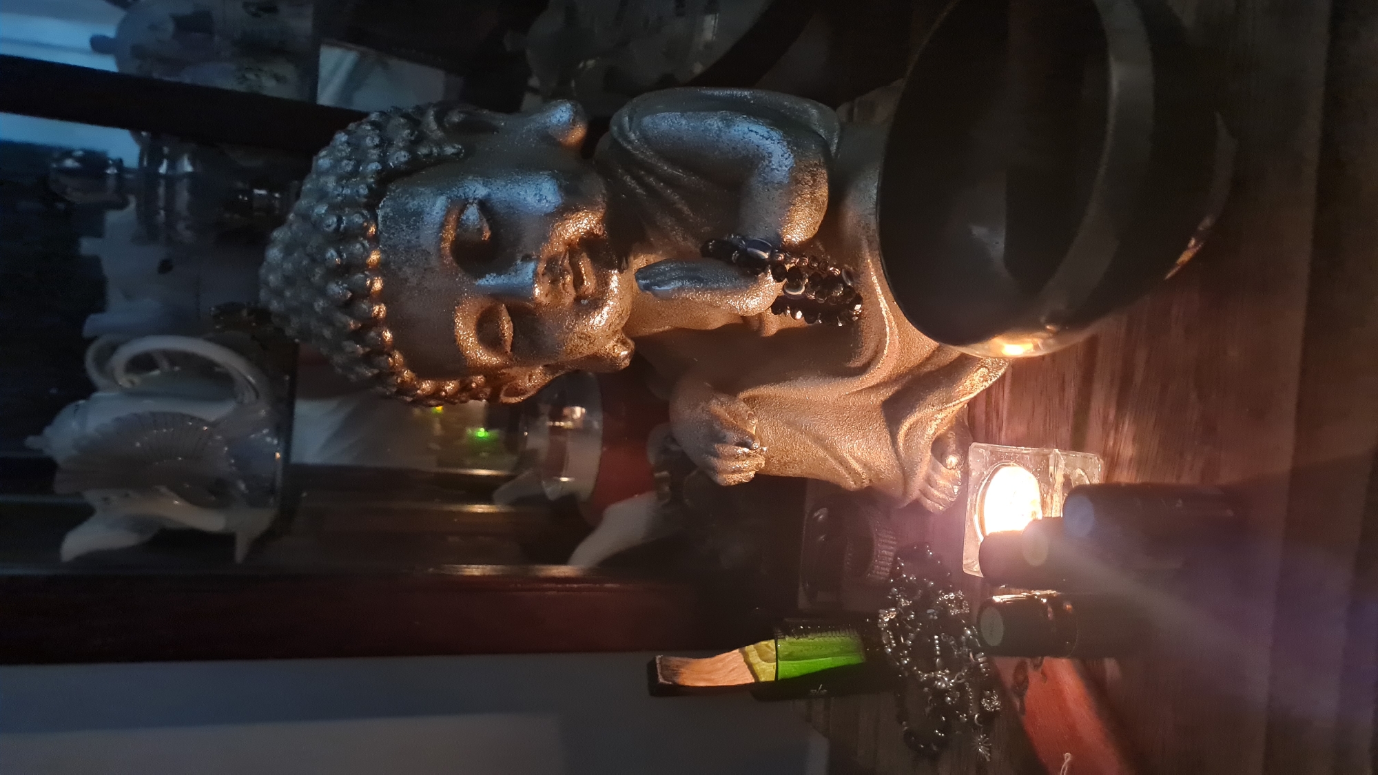 The image presents a still life arrangement. Dominating the scene is a Buddha statue, likely made of a metallic material, placed alongside a dark metal singing bowl. A lit candle in a square glass holder provides a warm light source, casting shadows and illuminating the arrangement. A black beaded necklace is draped around the base of the statue and near the candle. Additional items visible include a small green bottle and what appears to be a white porcelain swan figurine. The composition suggests a spiritual or meditative theme. Although not explicitly stated in the image, the user requested the image to be taken in Nahariyya, Israel.