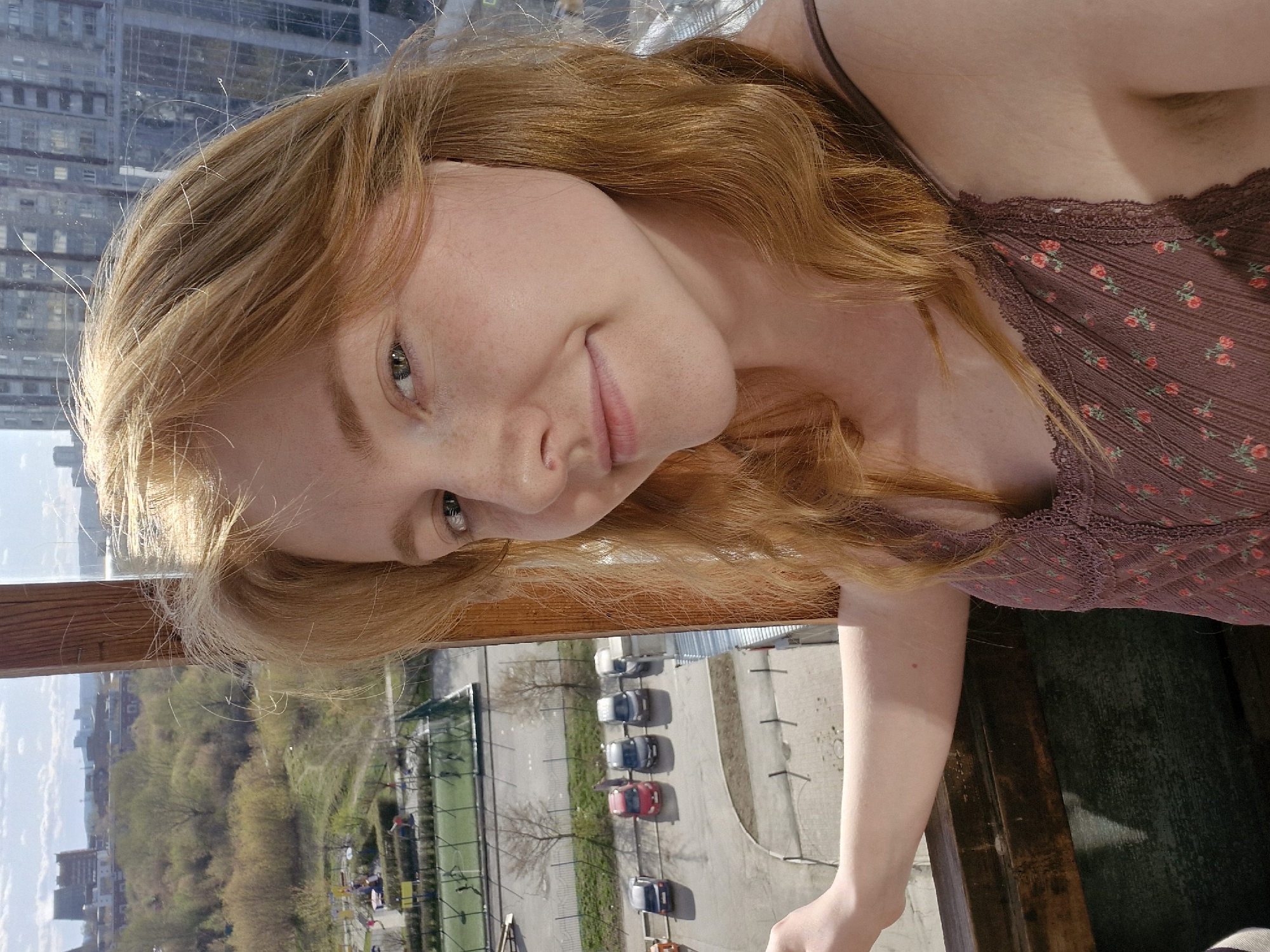 The image captures a young woman with long, wavy, reddish-blonde hair, her head tilted playfully as she looks towards the viewer with a gentle, slightly shy smile. Her green or hazel eyes are prominent, and faint freckles are visible across her nose and cheeks. She is dressed in a dark plum or maroon camisole, featuring delicate lace trim along the neckline and small, scattered floral patterns in shades of red and pink. Her left arm, pale and with a visible mole, rests casually on a sturdy wooden railing in the foreground.
The scene appears to be taken from a high vantage point, likely a balcony or an upper-floor window, offering an expansive view of an urban landscape. In the background behind her head, a tall, grey multi-story building with numerous windows indicates a densely built city area. Below the wooden railing, a paved parking lot stretches out, occupied by several parked cars of various colors. Adjacent to the parking lot is a green sports court, possibly for tennis, enclosed by fencing. Beyond these structures, a line of trees with fresh, light green foliage suggests spring or early summer. The sky above is clear and blue, dotted with a few wispy clouds, indicating a bright, sunny day. The overall impression is one of calm observation or a relaxed pose within a bustling city environment, consistent with a location such as Yekaterinburg, Russia. No visible text is present in the image.