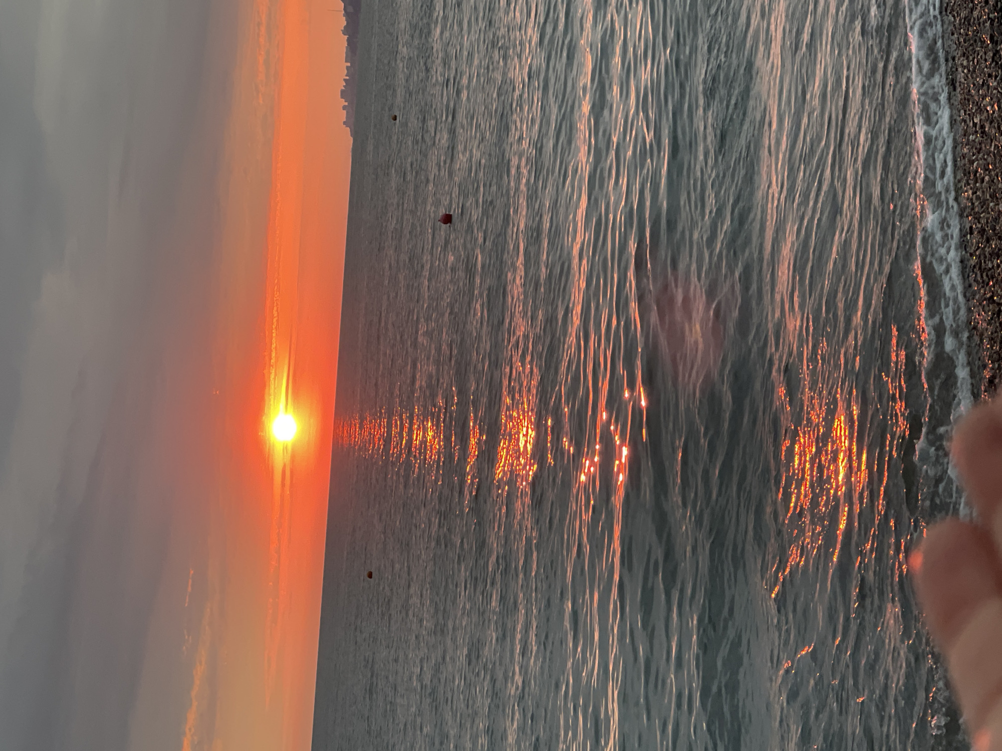 This is a vertical, eye-level media file of a sunset over the sea. The sun, a bright orange orb, is low on the horizon, casting a vibrant orange glow across the sky and reflecting on the water's surface as a shimmering path of light. The water is calm, with gentle waves creating ripples that catch the sunlight. In the foreground, to the right, a portion of a person's hand is visible, suggesting the photo was taken by someone standing at the edge of the water. The background on the left shows a hazy sky transitioning from orange to gray, with a faint silhouette of buildings on the distant shoreline, indicating an urban or coastal setting. On the water, two small, dark buoys are visible. The scene evokes a sense of tranquility and natural beauty. The lighting and colors suggest it is late evening, during sunset. The location is identified as Sochi, Russia.