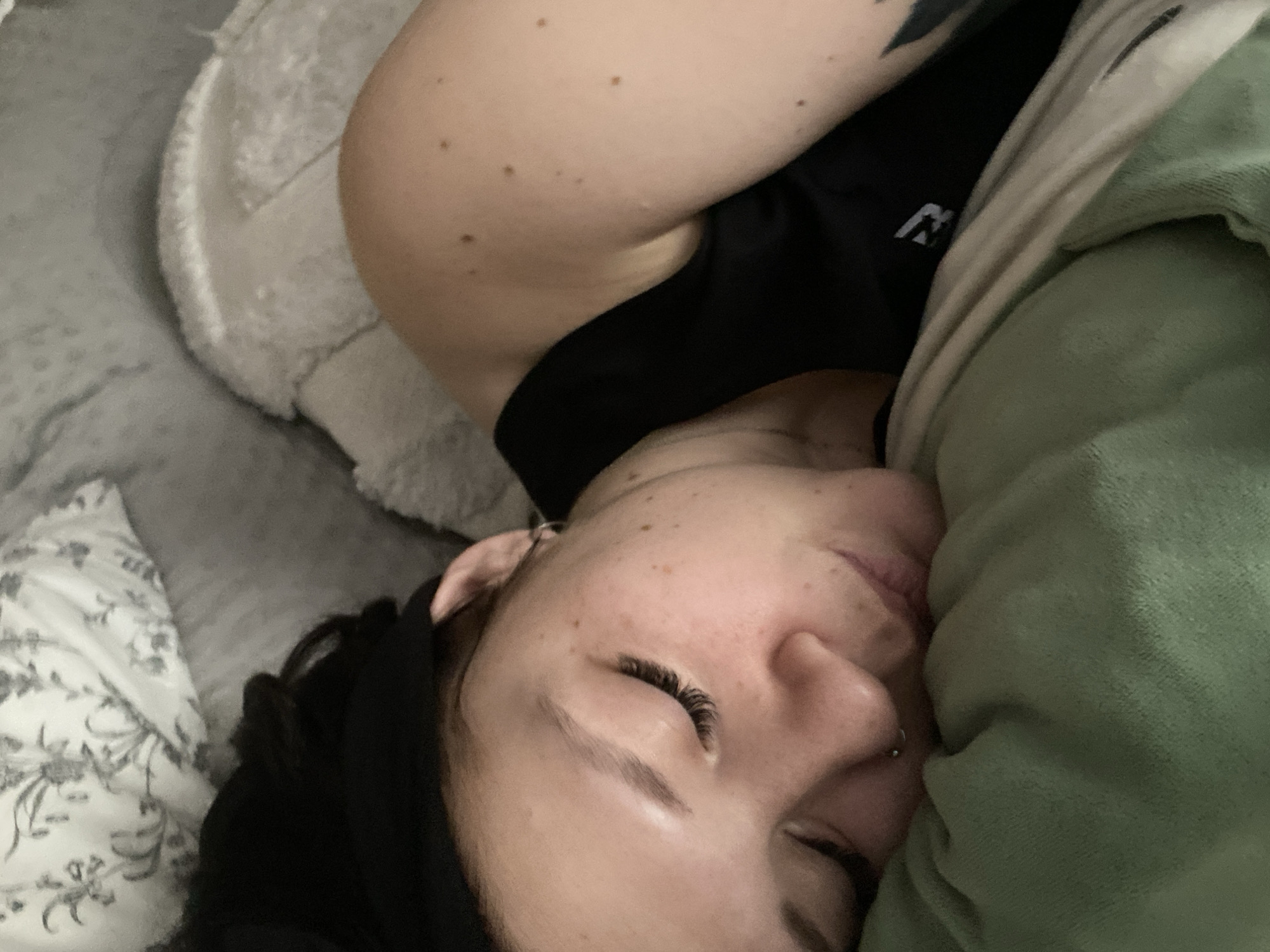 A person is lying in bed, possibly asleep. The person has dark hair and fair skin with freckles. They have pierced nostrils and wear eyelash extensions. A green blanket is pulled up close to their face. The person is wearing a black tank top. The bed has a floral-patterned pillowcase and a gray blanket. A white pillow is behind the person's head. A portion of an arm with moles and a tattoo is visible. There is no information in the image to indicate it was taken in Chaikovsky, Russia.