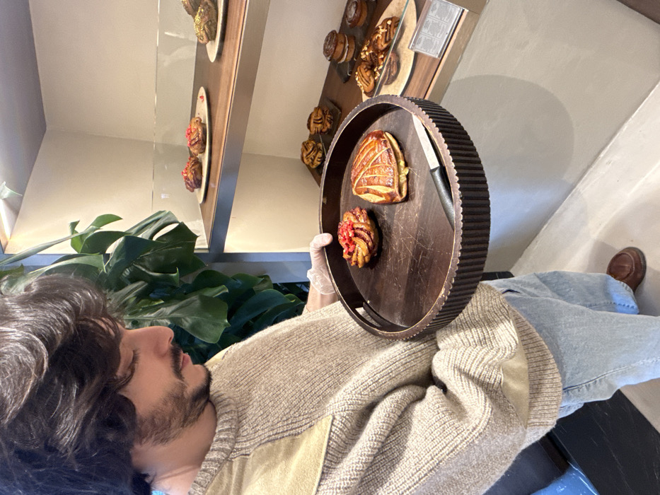The image depicts an indoor scene within a modern patisserie or bakery, likely situated in Paris, France. A male subject is positioned in the foreground, oriented in profile facing right. He possesses dark, wavy hair and a dark beard and mustache. His attire includes a beige knitted sweater with lighter beige patches on the shoulders, light blue jeans, and brown loafer-style shoes. A clear plastic glove covers his right hand.
The subject holds a round, dark brown wooden serving tray with a ribbed outer edge. On the tray are two distinct pastries: one spiral-shaped pastry topped with small red fruit pieces, and one golden-brown, crescent-shaped flaky pastry. A small knife with a black handle and a silver blade rests between the pastries.
In the background, a multi-tiered display case, featuring wooden shelves and glass partitions, showcases an assortment of baked goods. The top shelf holds plates containing spiral pastries, some adorned with red fruit, others with a green topping. Lower shelves display various other pastries, including multiple spiral-shaped buns and cylindrical stacked items, some topped with nuts. A small informational sign is visible on the bottom shelf. To the left of the display, a large green tropical plant, consistent with a Monstera species, is present. The interior environment features light-colored walls and a light-colored floor.