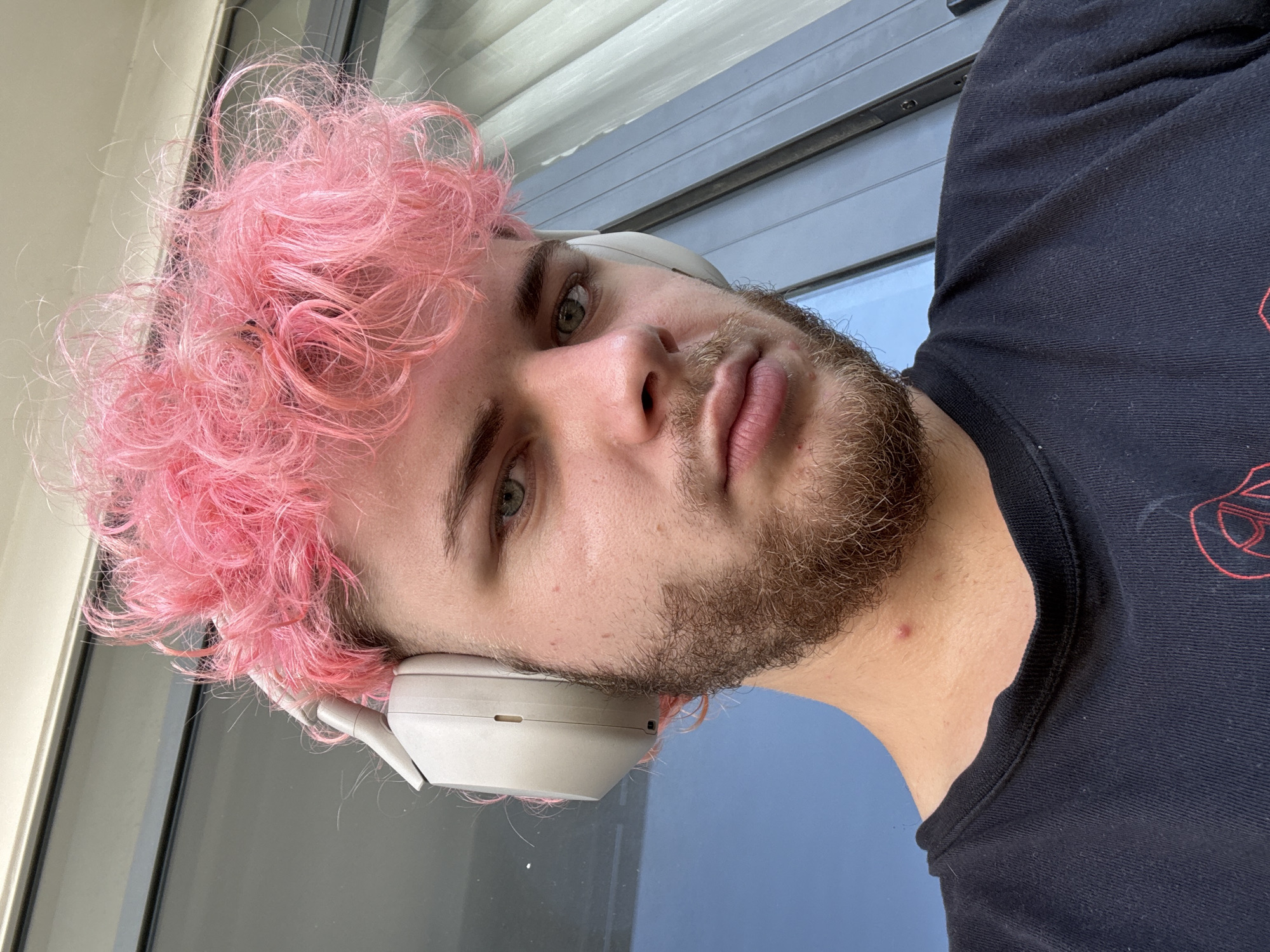 A close-up selfie photograph of a Caucasian male with pink curly hair wearing white headphones. He is wearing a black t-shirt. The background is a window and what appears to be a wall, likely indoors. There is a blemish on his neck. The location is not discernible from the visual information available.