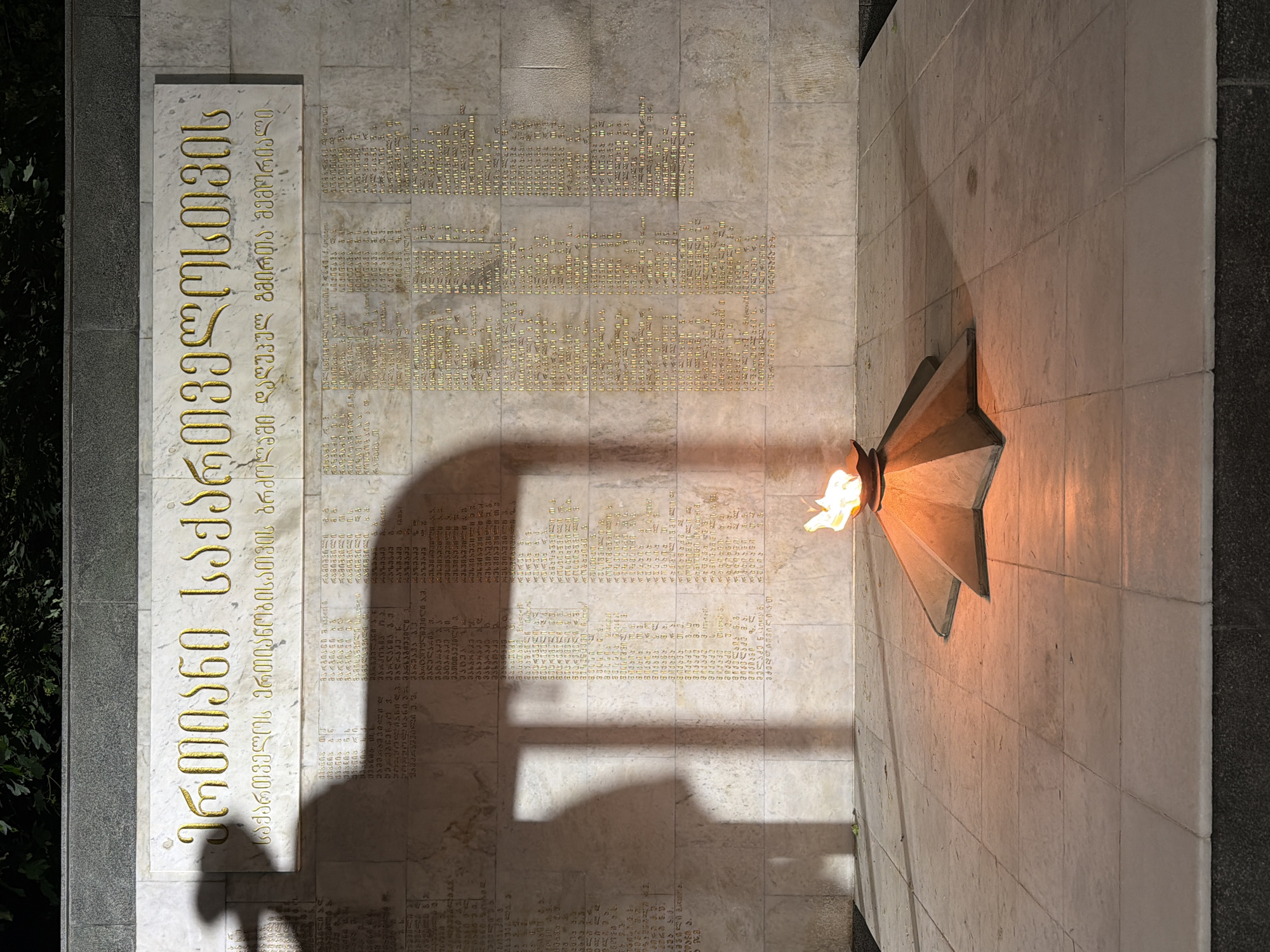 The image shows a memorial in Tbilisi, Georgia dedicated to those who died fighting for the unity of Georgia. A stone plaque on the left has golden Georgian script reading "For a Unified Georgia" and the inscription “A memorial to the heroes who died in the struggle for the unity of Georgia.” The wall behind the plaque has a grid of carved gold Georgian script. A five-pointed star-shaped structure on the right holds an eternal flame. A shadow falls across the wall. The surface of the wall is tiled with large rectangular stones.