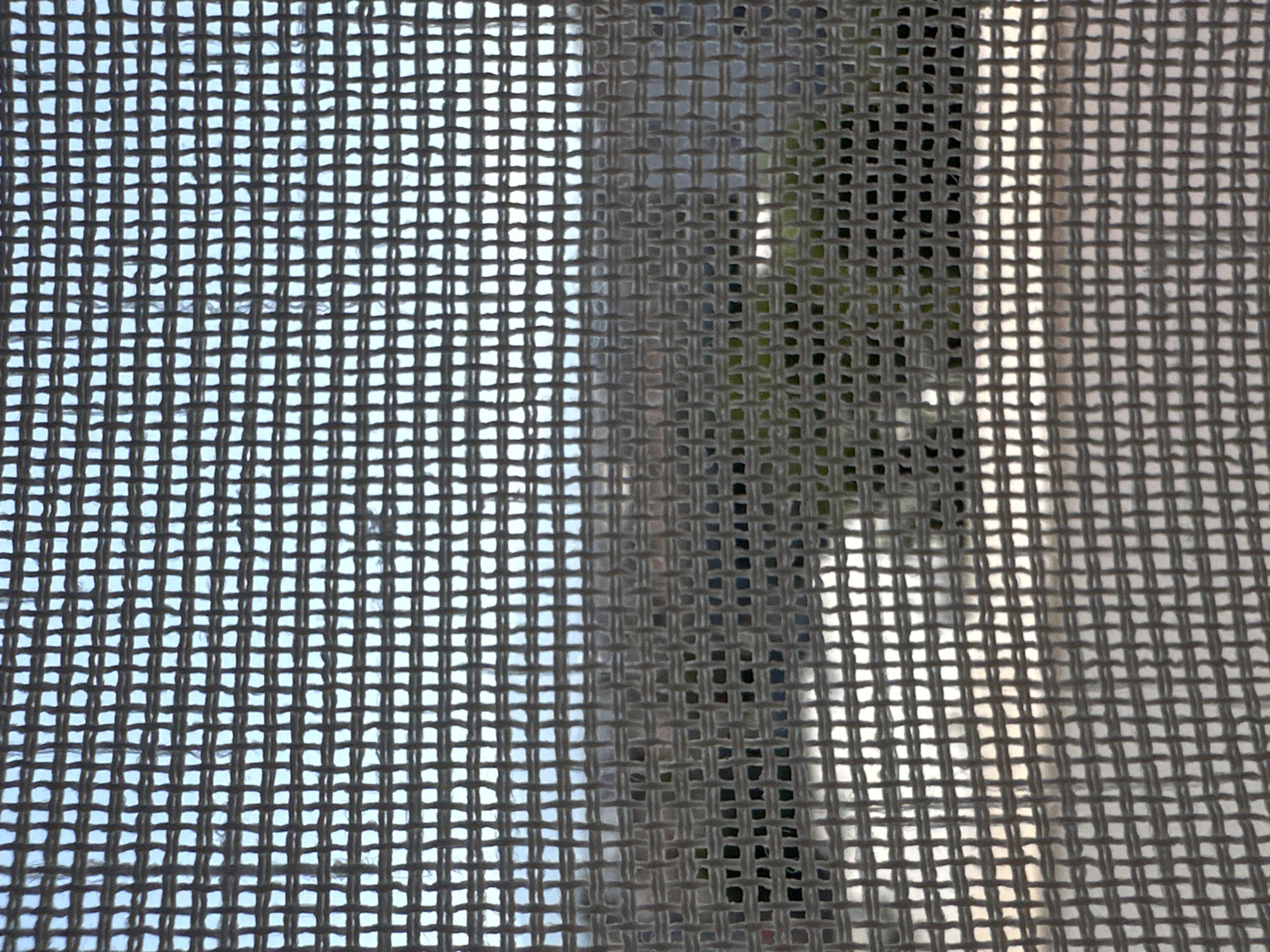 The image presents a close-up view of a window screen. The screen's mesh is clearly visible, with varying degrees of transparency that allow glimpses of the background. On the left, a lighter, blurry background suggests an open area with a blueish sky. The middle and right sides show a partially obscured view of what appears to be a building exterior, with rectangular shapes and possible greenery. Due to the limitations of the perspective, determining the exact location within Fethiye, Turkey, is impossible. There is no visible presence of people, interactions, or explicit actions within the confines of the frame.