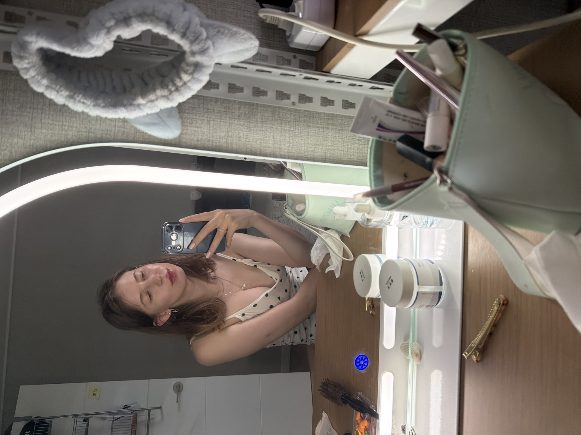 A young woman is taking a selfie in a mirror. She is wearing a white polka dot camisole and has brown hair. She is holding a phone in her right hand and is wearing a ring on her left hand. In the mirror's reflection, a shelf is visible with a grey fuzzy headband with ears. On the desk in front of the mirror are various makeup items, including jars, tubes, brushes, and a makeup bag. A hairbrush is also visible. The scene appears to be in a bedroom or dressing area. The location is identified as Dongducheon-si, South Korea.