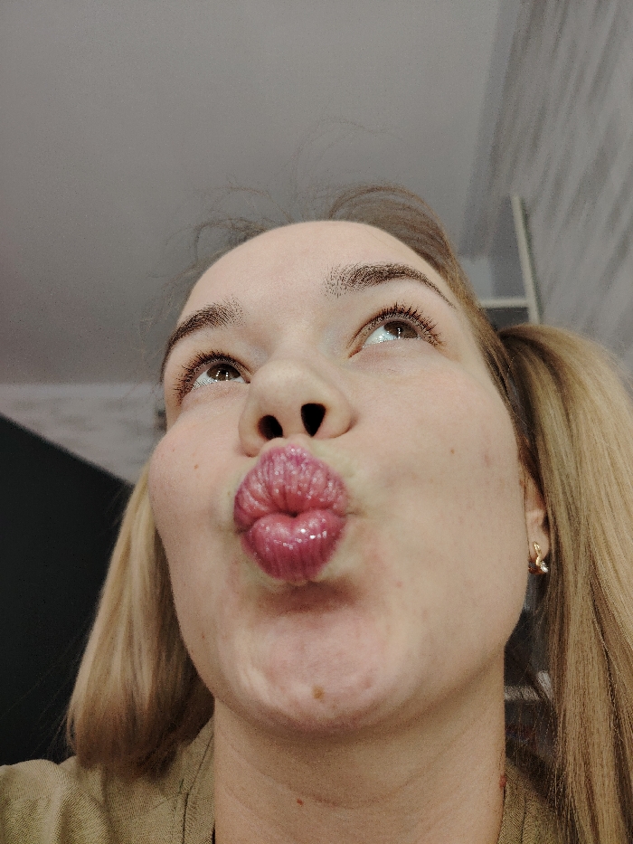 A woman with blonde hair pulled into pigtails puckers her glossy lips in a kiss. Her face is tilted upward, and her eyes gaze off to the side. She has visible freckles on her cheek and neck. She is wearing a gold earring. The background includes a glimpse of a dark object to the left and a white wall or shelf to the right.