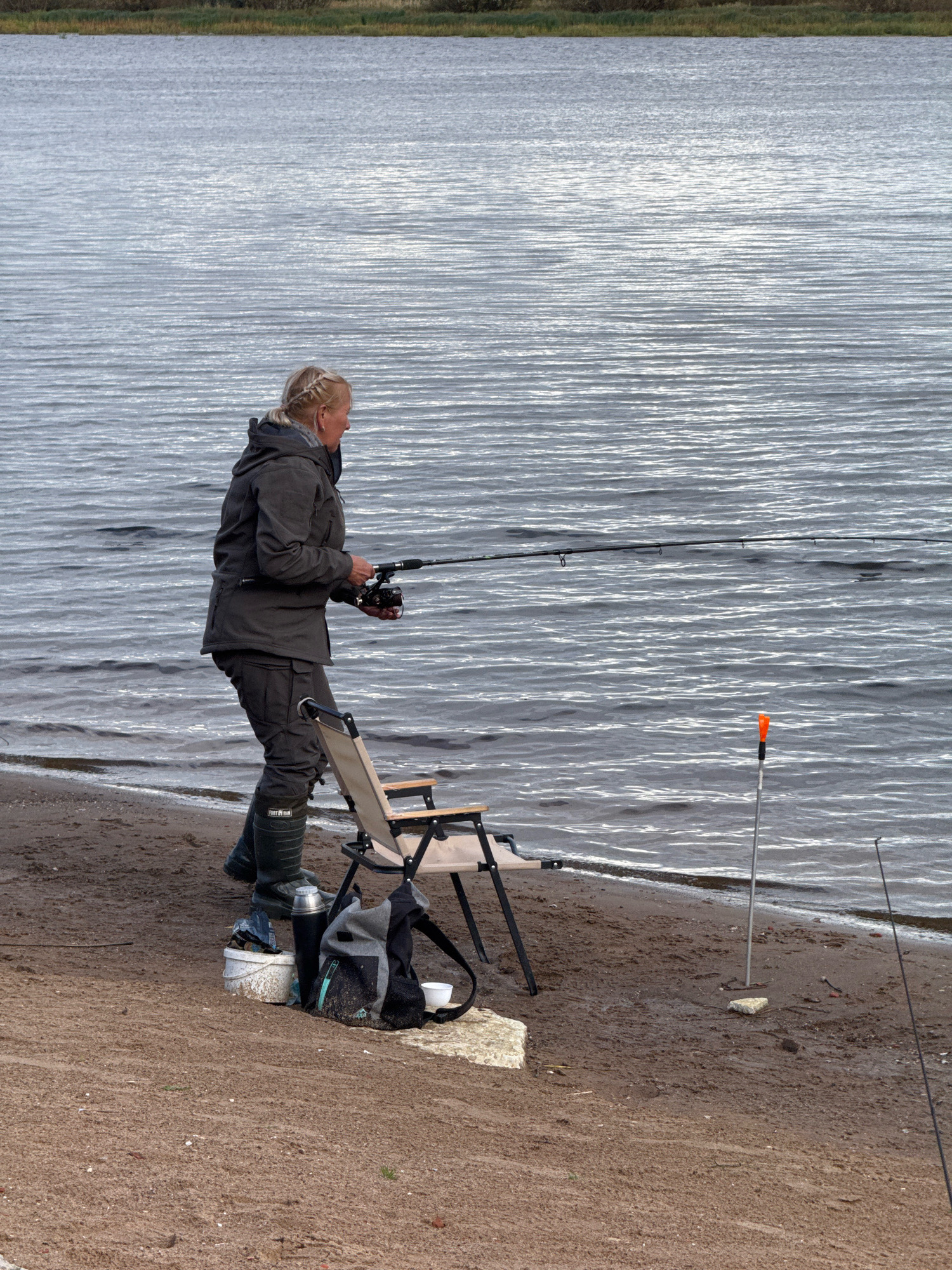 A woman is fishing on the shore of a lake, likely in Veliky Novgorod, Russia, given the context. She is wearing dark waterproof overalls and tall green rubber boots. She holds a fishing rod and reel in her hands, actively engaged in fishing. Beside her, a portable fishing chair is set up, along with a backpack, a white bucket, and a thermos. A fishing float is visible in the water near the shore. The environment consists of a sandy beach and a body of water with a rippled surface, indicating a moderate breeze. The sky is overcast.