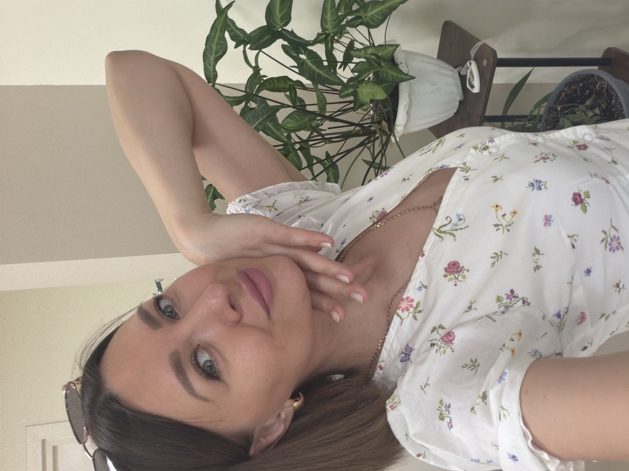 A selfie of a woman with brown hair is turned sideways. She is wearing a white dress with floral patterns and a gold necklace. Her left hand is raised to her face, with her fingers touching her cheek. Sunglasses are visible near her hair. In the background, a green plant sits on a shelf next to another potted plant. The location is implied to be an indoor setting in the city of Rostov-on-Don, Russia.