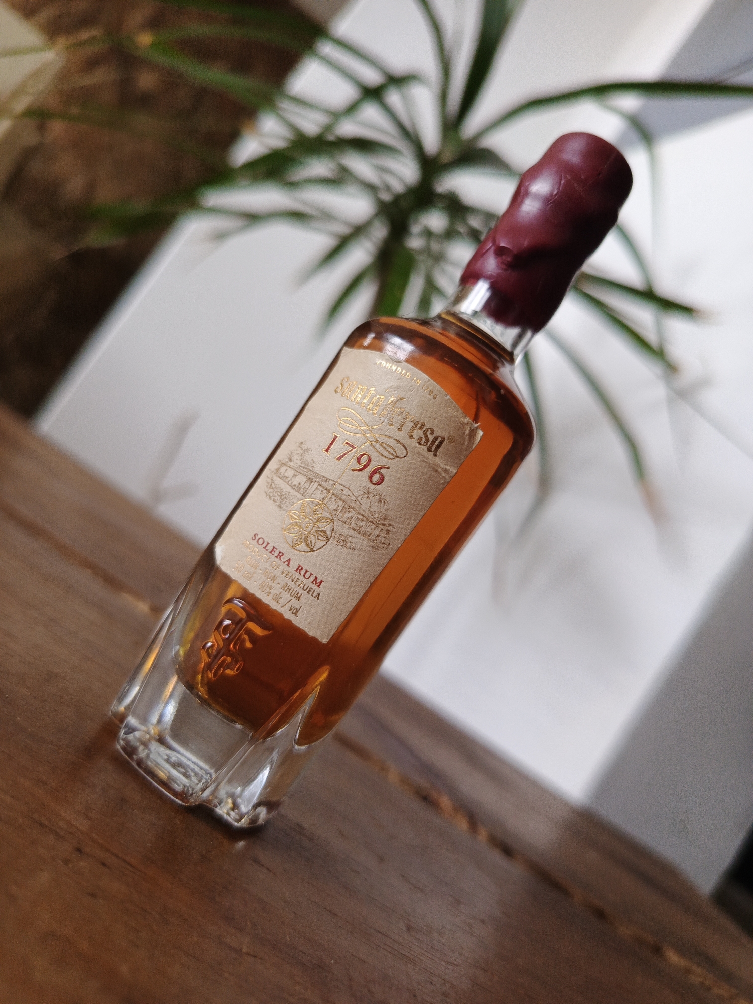 A slightly angled, medium shot depicts a bottle of Santa Teresa 1796 Solera Rum, situated on a wooden surface against a blurred backdrop of greenery and a white wall. The rum, bottled in a uniquely shaped clear glass, contains an amber-colored liquid. The bottle's label is beige with gold lettering, displaying the brand "Santa Teresa 1796" along with the words "SOLERA RUM," "PRODUCE OF VENEZUELA," and "RU-RON-RHUM." The bottle's neck is sealed with a maroon-colored wax. The wooden surface appears to be a table, and the setting suggests an indoor environment. The mention of Venezuela and Solera Rum alludes to production in the Los Robles region, but it is not specifically stated in the image.