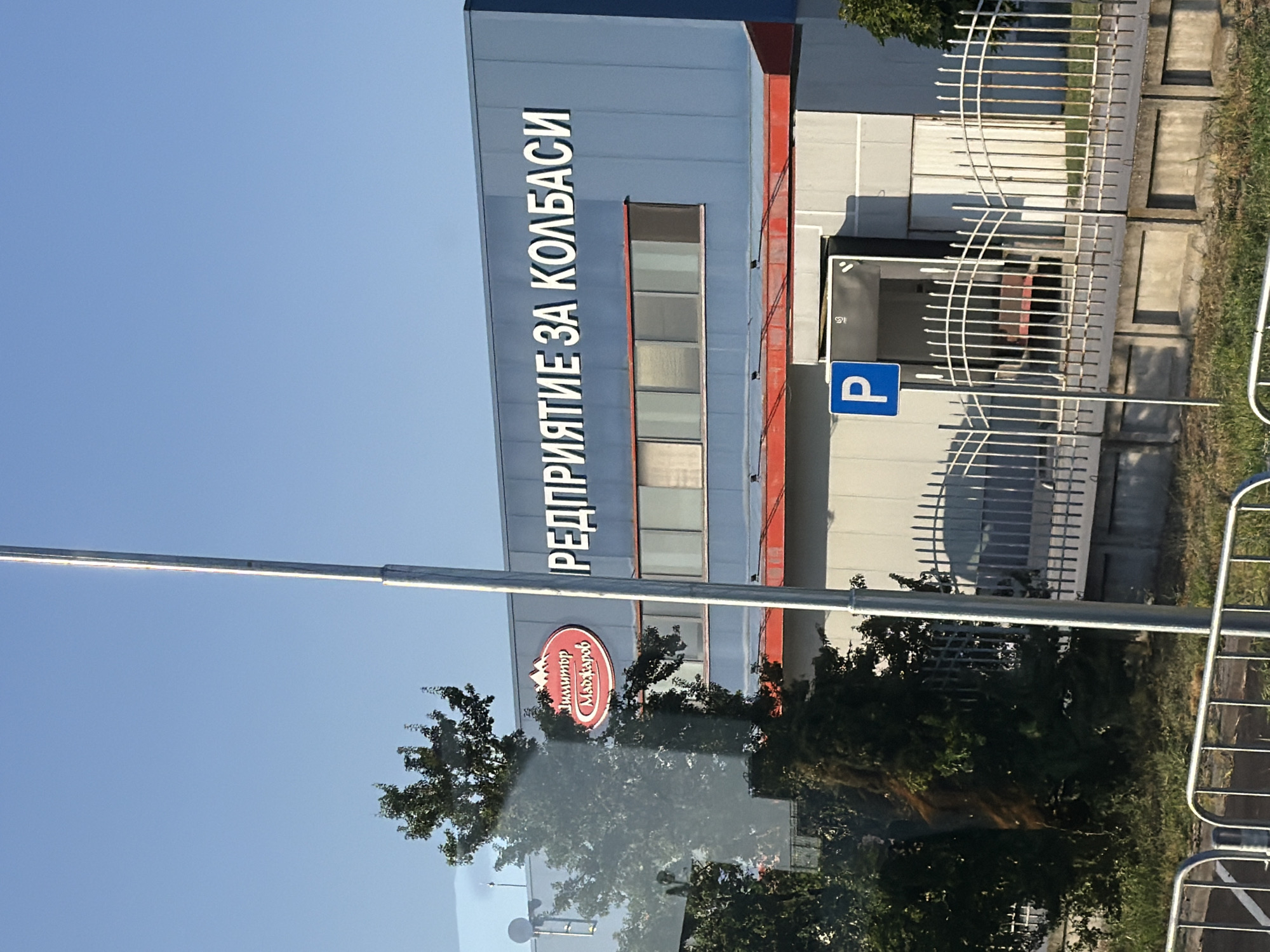 The image shows a building with signage that reads "РЕДПРИЯТИЕ ЗА КОЛБАСИ" which translates to "Sausage Enterprise." There is also a circular logo with mountains and the text "Димитър Маджаров" which appears to be a brand name. The building is blue with several windows. In front of the building, there is a white metal fence and a parking sign. Trees and grass are visible in the foreground and surrounding areas. The scene appears to be in an industrial or commercial area under a clear blue sky.