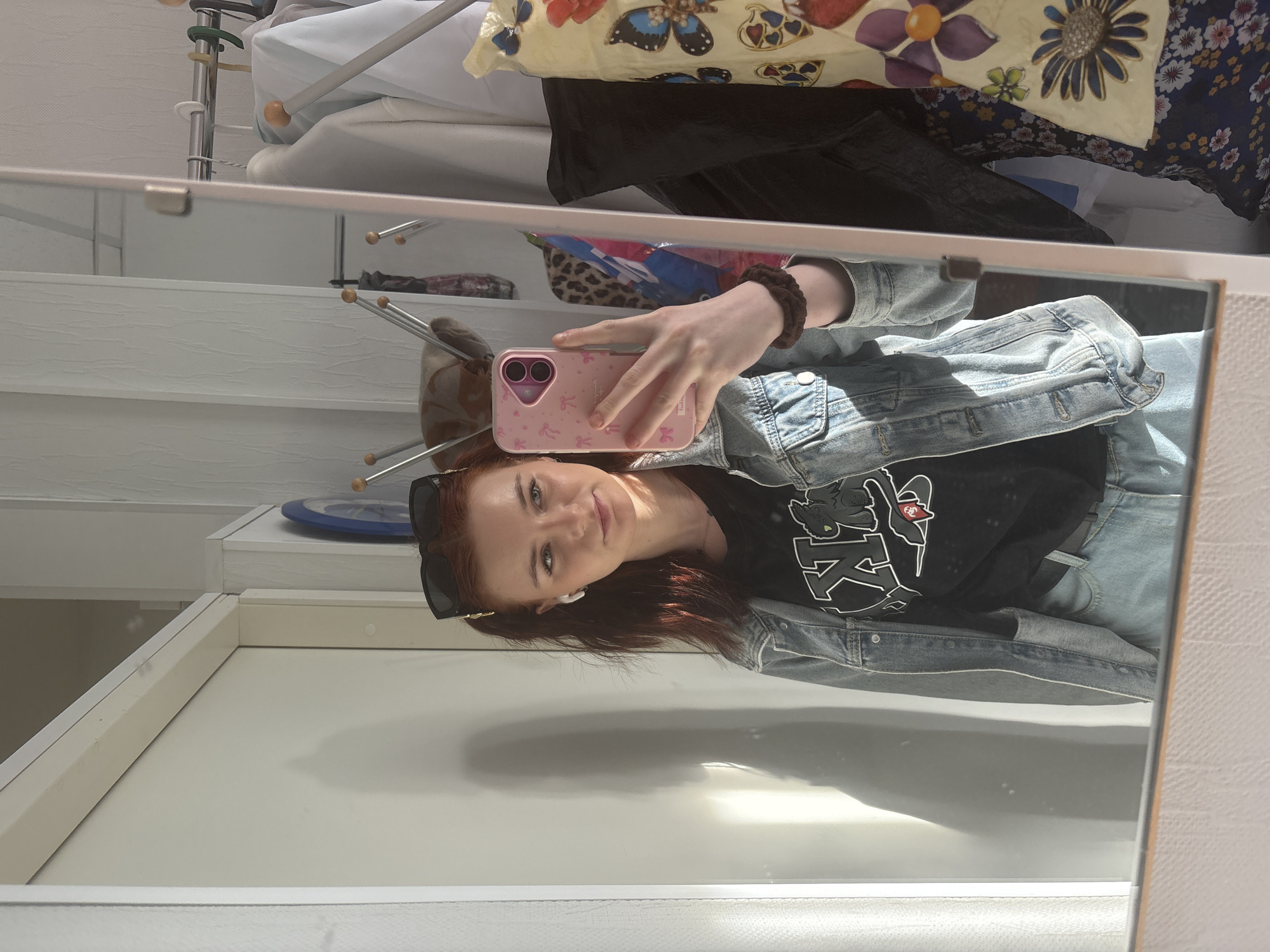 A young woman with auburn hair is taking a selfie in a mirror. She is wearing sunglasses on her head, a black t-shirt, and a denim jacket. Her phone is pink with a decorative case. Behind her is a white wardrobe with clothes hanging on a rack, including a floral-patterned item. The location appears to be an indoor space, possibly a room in Saint Petersburg, Russia, although no definitive indicators of the city are present in the image. The scene is brightly lit, with a clear reflection in the mirror.