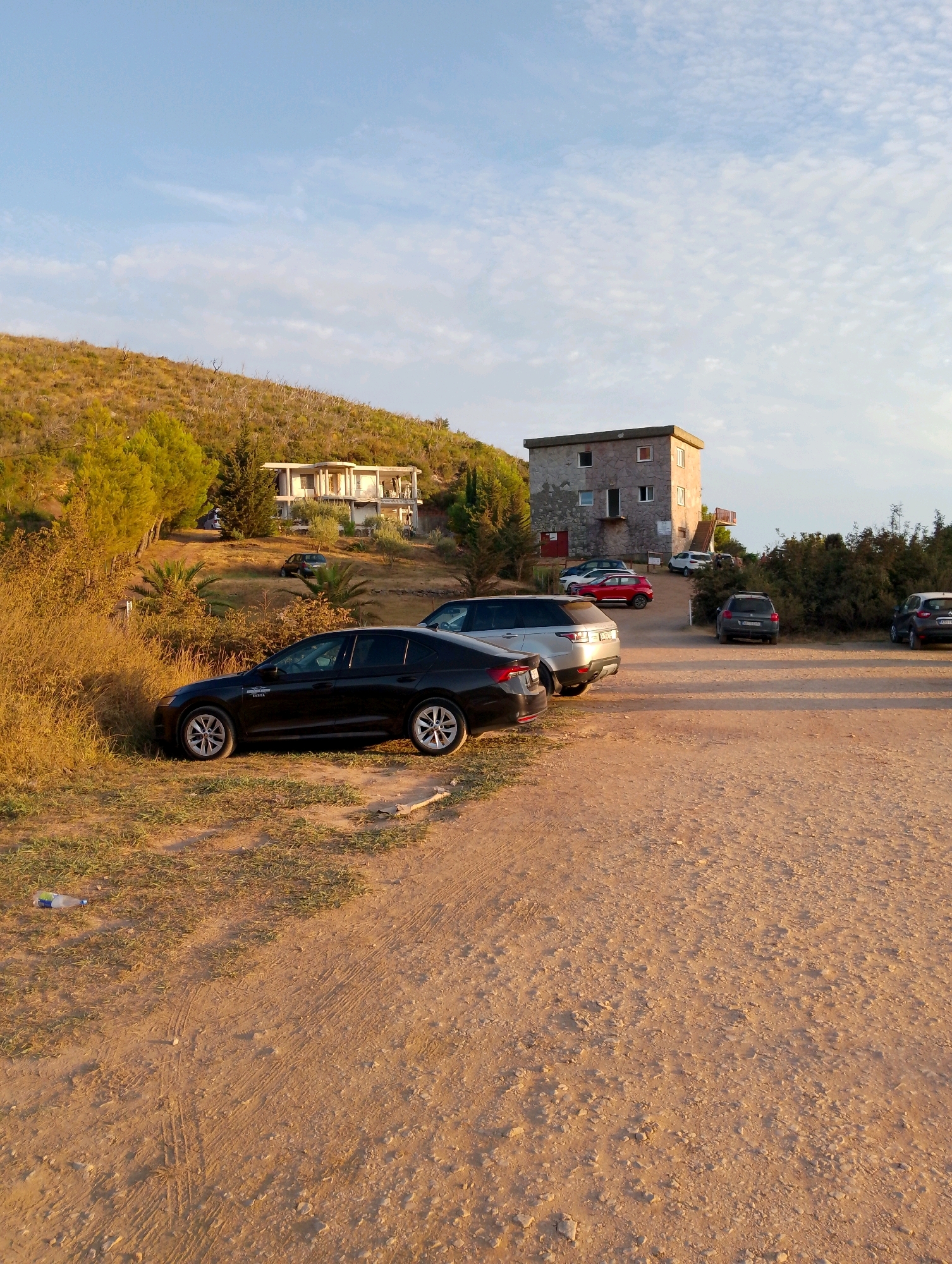 The image depicts a parking area with several vehicles on a dirt surface, likely located in Sutomore, Montenegro. In the foreground, a black sedan is parked on the left side of the frame, partially on dry grass. Further back, a silver SUV is parked, followed by a dark grey SUV. To the right, two other vehicles are visible. Behind the parking area, a two-story concrete building with multiple windows and an external staircase stands on a hillside. The hillside is covered with dry vegetation and some scattered trees. The sky is partly cloudy with a light blue hue, suggesting it is either morning or late afternoon. A plastic bottle lies on the ground near the black sedan. The overall scene is dry and sun-drenched, characteristic of a Mediterranean climate.