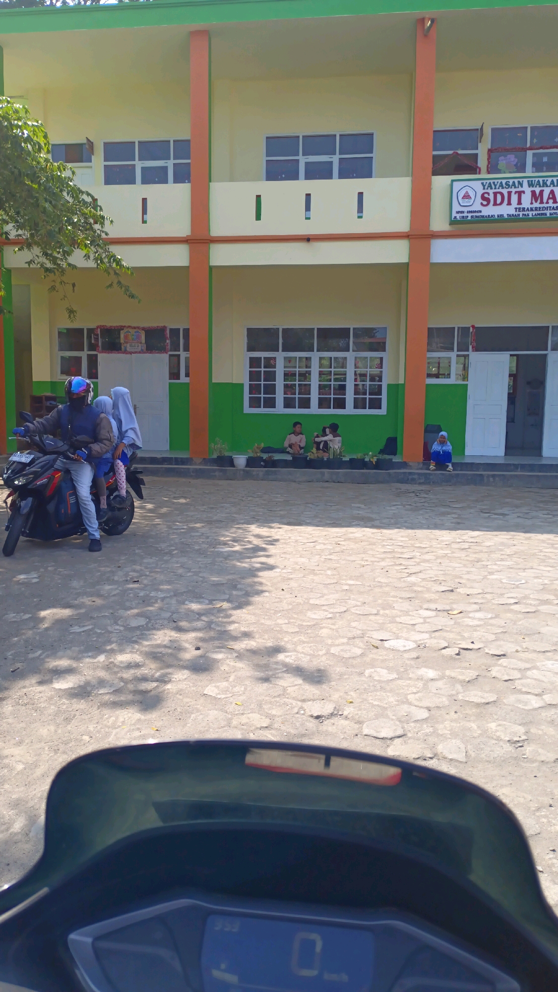 A person rides a motorcycle with two passengers in front of a school building in Padang Panjang, Indonesia. The school building is a two-story structure painted yellow and green with orange columns. A sign at the entrance reads "YAYASAN WAKAF" and "SDIT MA". Several students are seated on the steps of the school. The ground is paved with concrete blocks, and there are shadows cast by trees. The motorcycle is black and red, and the passengers are wearing casual clothing, with one child wearing a hijab. The image is taken from the perspective of someone on another motorcycle, showing a partial view of its handlebars and dashboard.