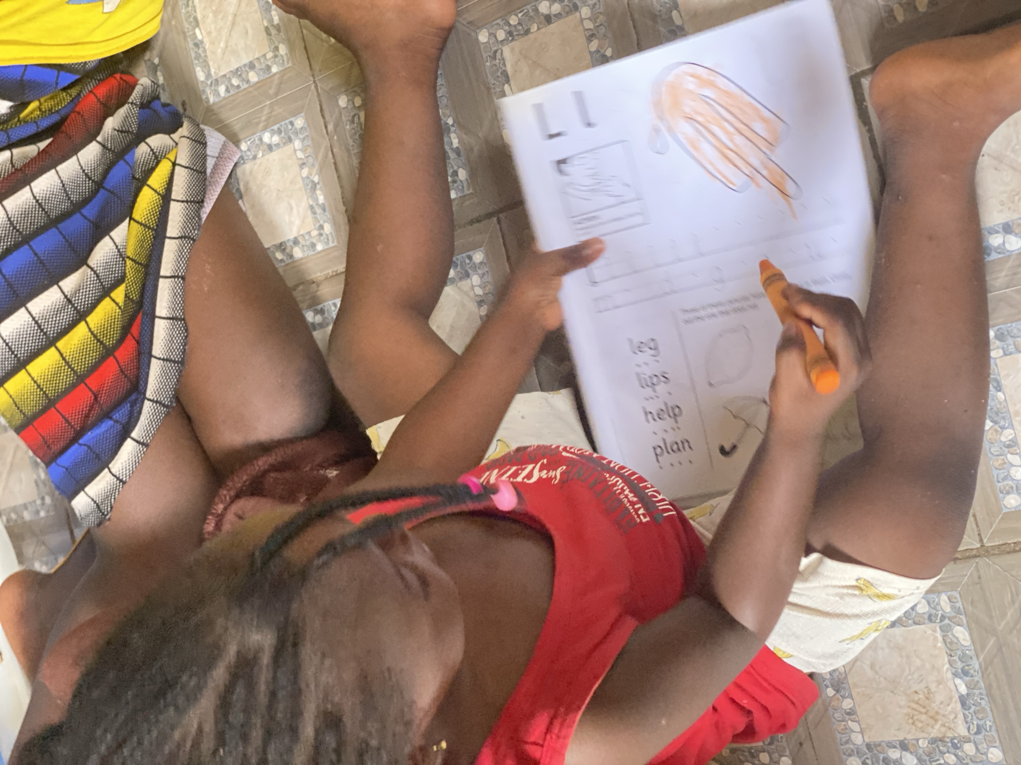 A child is sitting on a tiled floor in Rido, Nigeria, coloring a worksheet with an orange crayon. The worksheet features the letter "L" and illustrations of a hand, leg, lips, and an umbrella, alongside corresponding words. The child wears a red shirt and is positioned between the legs of an adult wearing shorts, one pair multicolored and one pair patterned with small yellow birds.