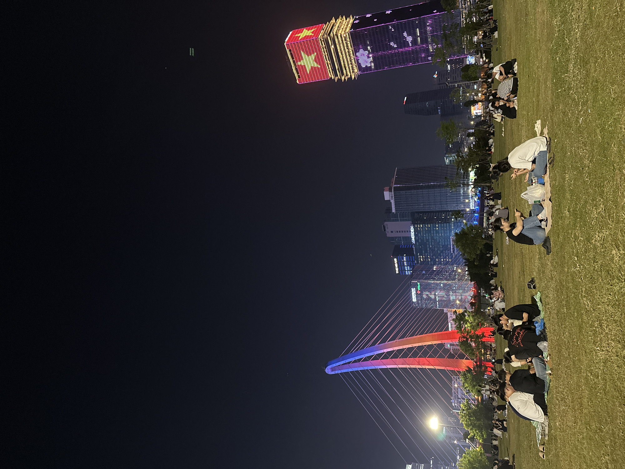 Nighttime scene in Thủ Đức, Vietnam. People are gathered on a grassy area, seemingly a park. Buildings are visible in the background. One building is illuminated with the Vietnam flag. A cable-stayed bridge, lit with red and blue, is in view. People are sitting on blankets and interacting.