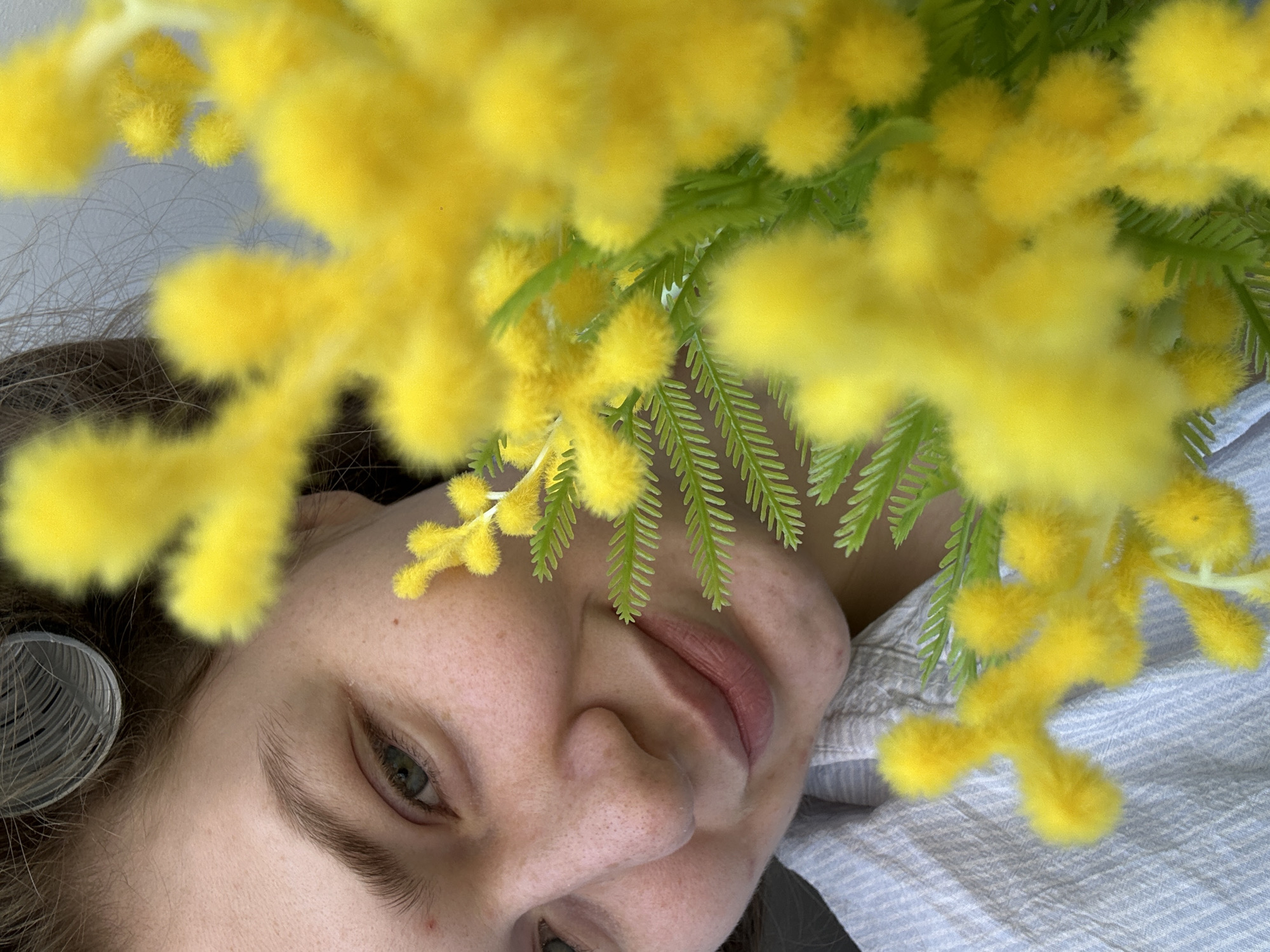 A close-up, slightly angled, portrait features a fair-skinned woman partially obscured by sprigs of yellow mimosa flowers with green feathery leaves. The woman's face is the primary focus, revealing an eye with brown lashes, a nose, and slightly parted lips. A hair curler is visible in her hair near her left eye. The background is a light-colored, striped fabric. The presence of mimosa may indicate a celebration of International Women's Day, but no details in the image confirm this. The image provides no information to identify its location as Kazan, Russia, or any other city.