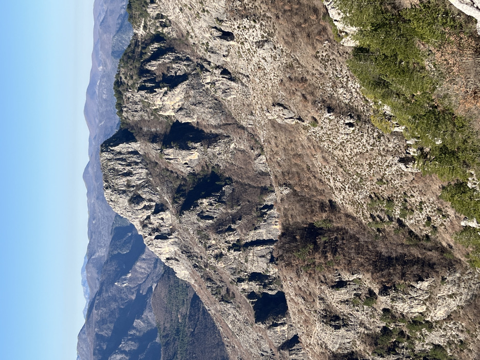 The image depicts a vast, rugged mountainous landscape under a clear, pale blue sky, located in the vicinity of Klobuk, Bosnia and Herzegovina. The foreground and midground feature steep, rocky slopes characterized by exposed gray and brown rock formations, interspersed with sparse vegetation. Patches of evergreen trees, primarily pine, appear in the lower right and along some ridges, contrasting with areas of deciduous growth that are largely bare or possess minimal brown foliage. A prominent, jagged rock peak dominates the mid-left, exhibiting significant erosion and vertical fissures. Multiple layers of mountains extend into the background, becoming progressively hazier and bluer, indicating considerable distance. Sunlight illuminates the scene, casting shadows that accentuate the three-dimensional topography of the terrain. No human activity, structures, or animals are visible.