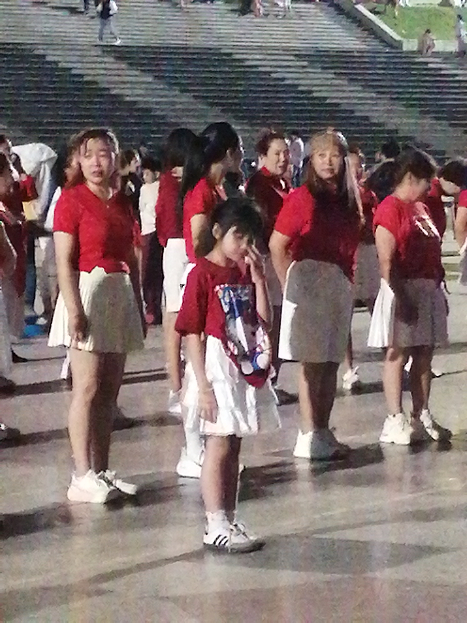 The image shows a group of people, mostly women and a young girl, gathered outdoors. They are wearing red tops and white skirts, suggesting a performance or uniform. The setting appears to be a public space with tiered seating in the background, reminiscent of an outdoor amphitheater or grandstand. The ground is paved and reflects light, indicating it might be damp or polished.
The group seems to be participating in an event, possibly a dance performance or a coordinated activity. The young girl in the foreground is looking towards the camera, and her hand is raised to her face. The other individuals are facing different directions, some looking towards the front, others engaged with their surroundings.
The lighting suggests it could be late afternoon or early evening, as there are shadows cast on the ground. The weather seems clear. There are no discernible signs or text in the image. The overall mood appears to be casual and perhaps a bit formal due to the matching outfits. The presence of stairs and open space suggests a public gathering place. The context of Da Lat, Vietnam, places this scene in a Southeast Asian urban or park environment.