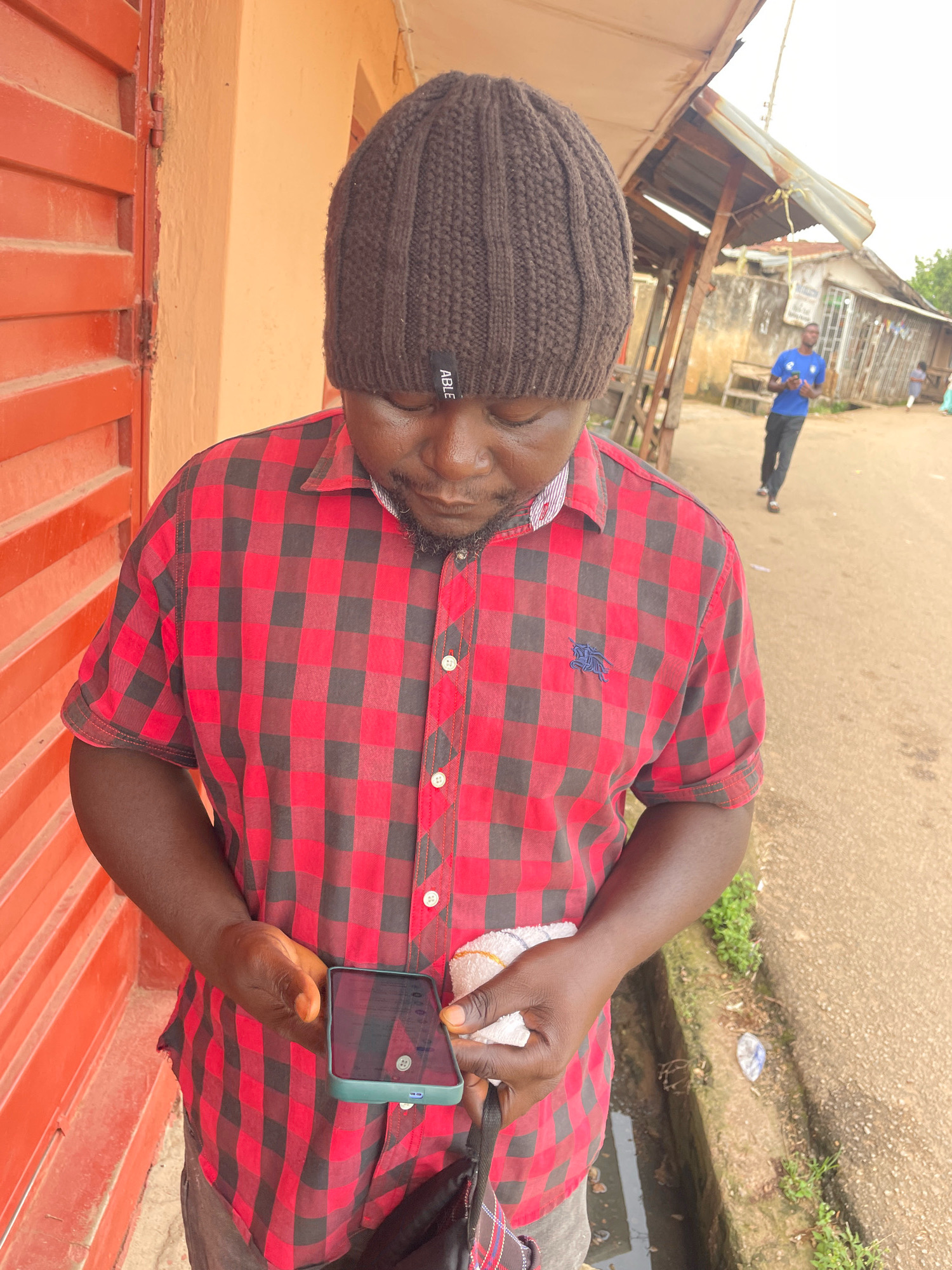 In Jos, Nigeria, a man wearing a brown knitted beanie and a red and black checkered short-sleeved shirt stands in front of an orange-red corrugated metal door. He is looking down at a smartphone he holds in his left hand. In his right hand, he holds a white cloth. In the background, a dirt road is visible, with a few buildings lining the side and a person in a blue t-shirt walking away from the camera. The area appears to be an outdoor market or street scene.