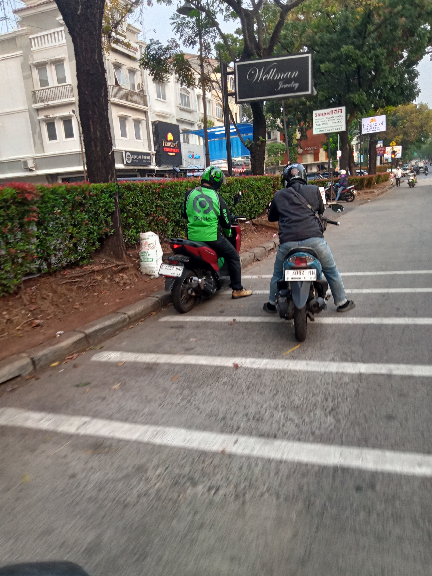 In South Tangerang, Indonesia, on a street with pedestrian crossing markings, two individuals on motorcycles are captured in motion. The motorcycle on the right is being ridden by a person wearing a black jacket and blue jeans, with a black helmet. The motorcycle on the left is ridden by a person in a bright green jacket and black pants, also wearing a helmet. The background features a multi-story building with balconies, a tree with dense foliage, and various signs, including "Wellman Jewelry," "SIMPELL JR BADAN USAHA," and "House of Croissant." A white sack with text that is partially obscured is visible on the side of the road near the left motorcycle. The scene is indicative of urban street traffic.