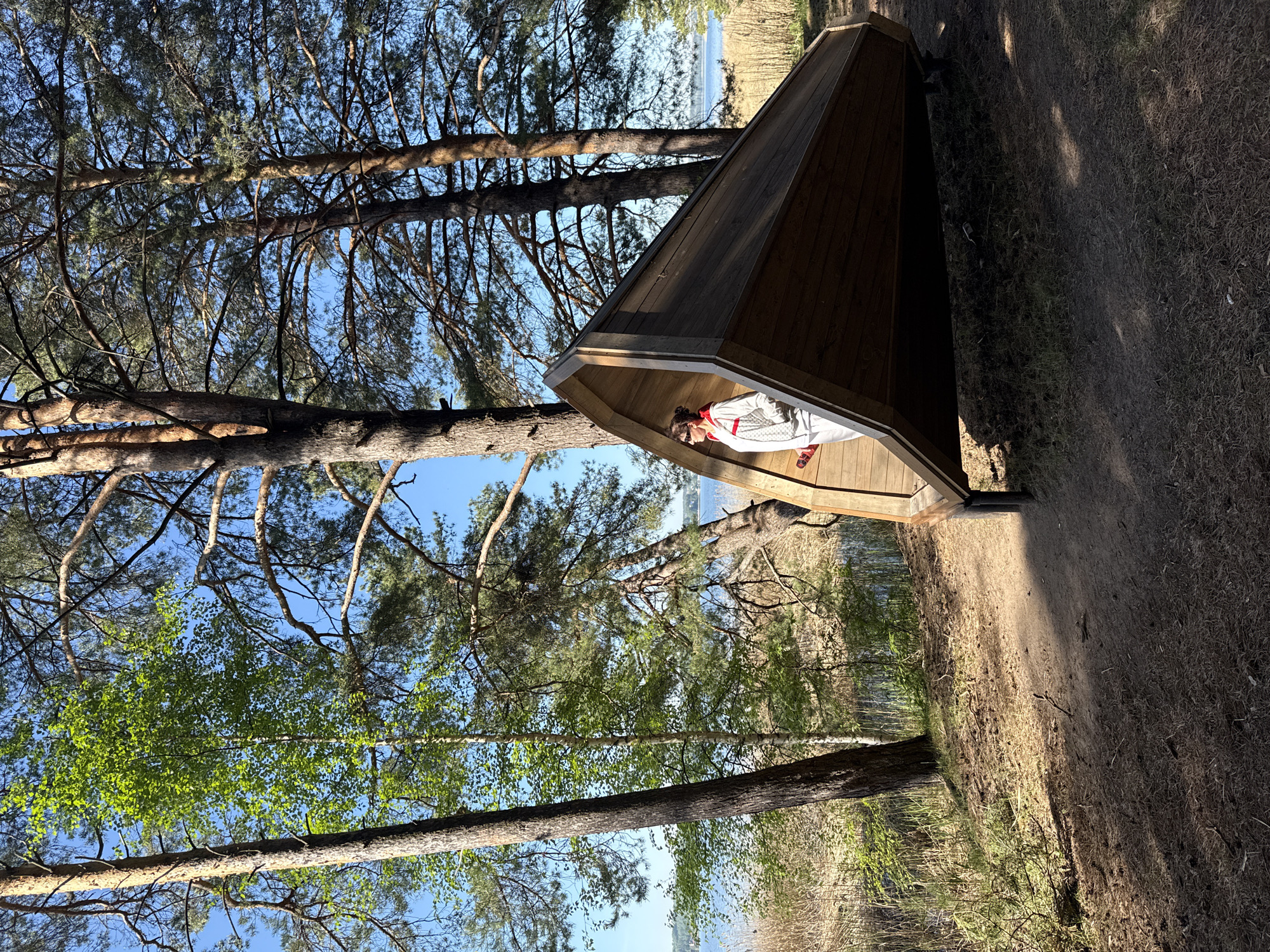 The image depicts an outdoor scene in Votkinsk, Russia. A wooden structure, resembling a small hut with a pitched roof, is positioned amidst trees and vegetation. Inside the structure, a person is visible wearing a white outfit with red accents. The surrounding environment consists of trees, with some appearing to be pine trees. The ground is covered in grass and dirt. In the background, a body of water is partially visible through the trees. The lighting suggests it is daytime.