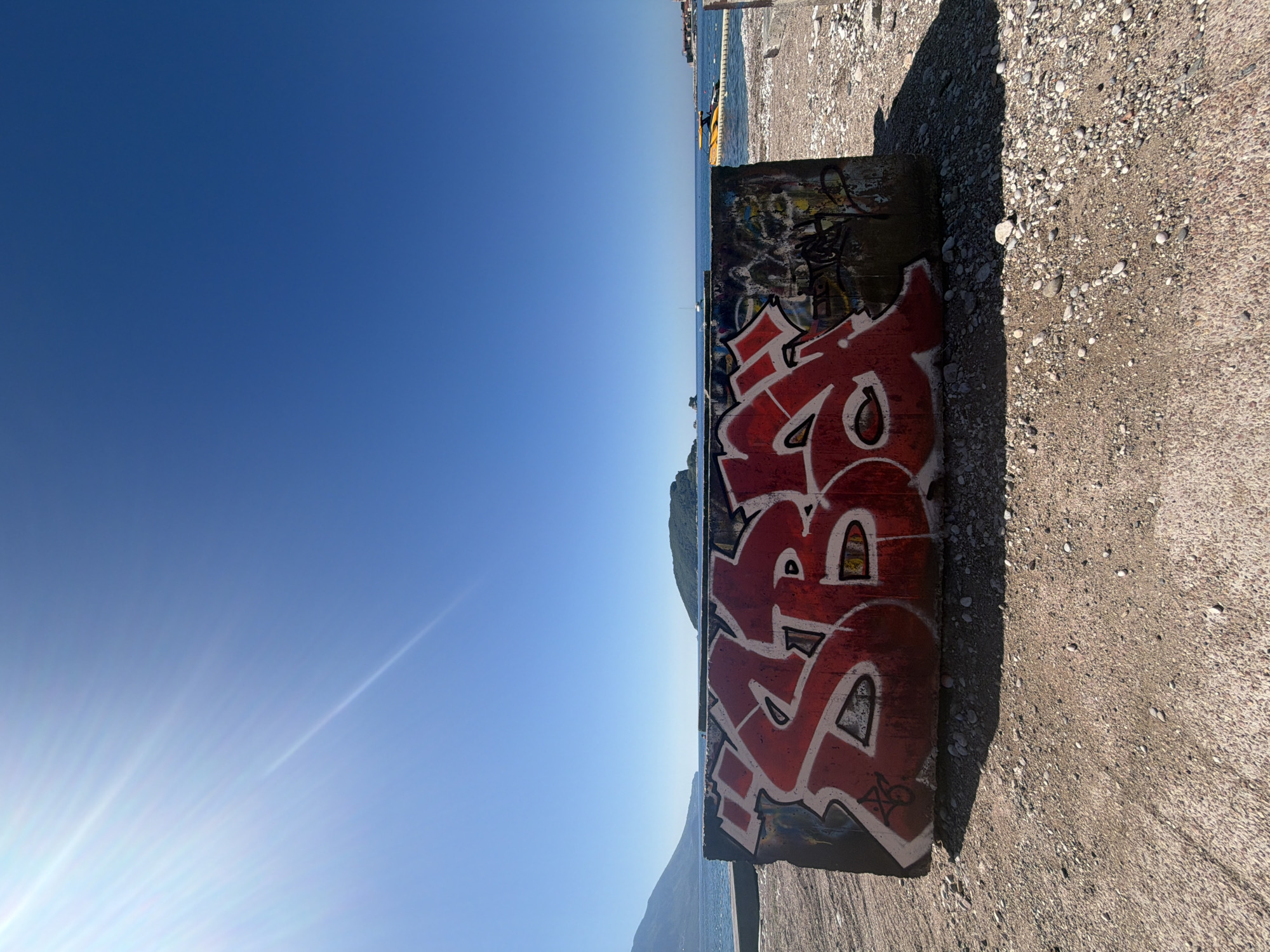 The image shows a concrete structure covered in graffiti. Red and white graffiti is most prominent, with some other darker, less defined graffiti near the top. The structure sits on a concrete surface with pebbles and other debris. To the left of the structure is the Adriatic Sea and a clear, blue sky. Across the water are mountains with some greenery. The location appears to be near the sea. Based on the presence of the sea and surrounding landscape, this scene is plausibly located in Budva, Montenegro.