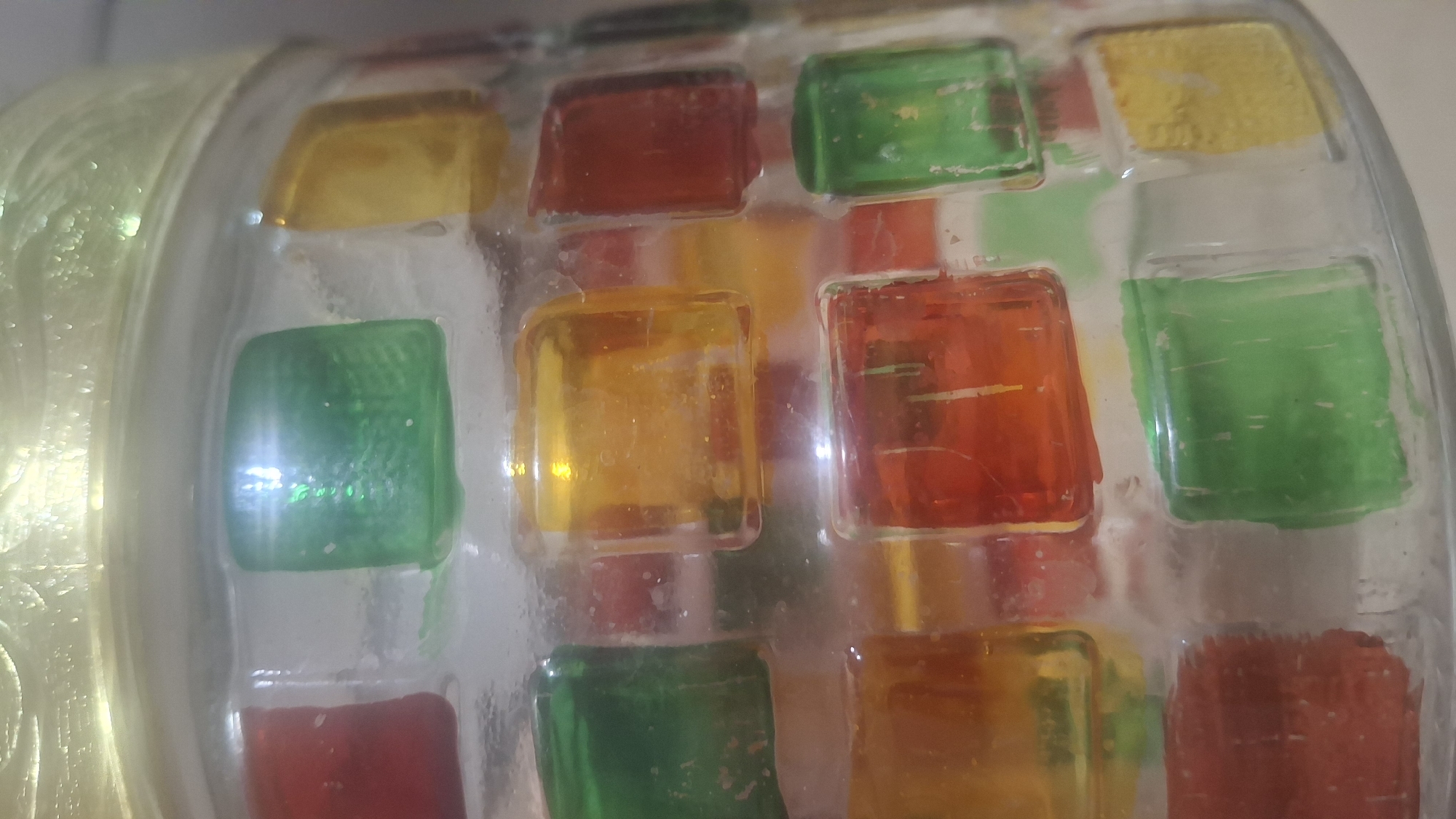 The image shows a close-up of a glass jar decorated with colorful, square mosaic tiles in red, green, and yellow. The tiles appear to be embedded in clear resin or glass, creating a textured and somewhat translucent surface. The jar has a golden, ridged lid that is partially visible at the top. The lighting suggests the jar is illuminated from above, creating highlights on the glossy surfaces of the tiles. There are no people or any discernible elements of the city of Myadzyel, Belarus visible in this image. The focus is entirely on the decorative jar.