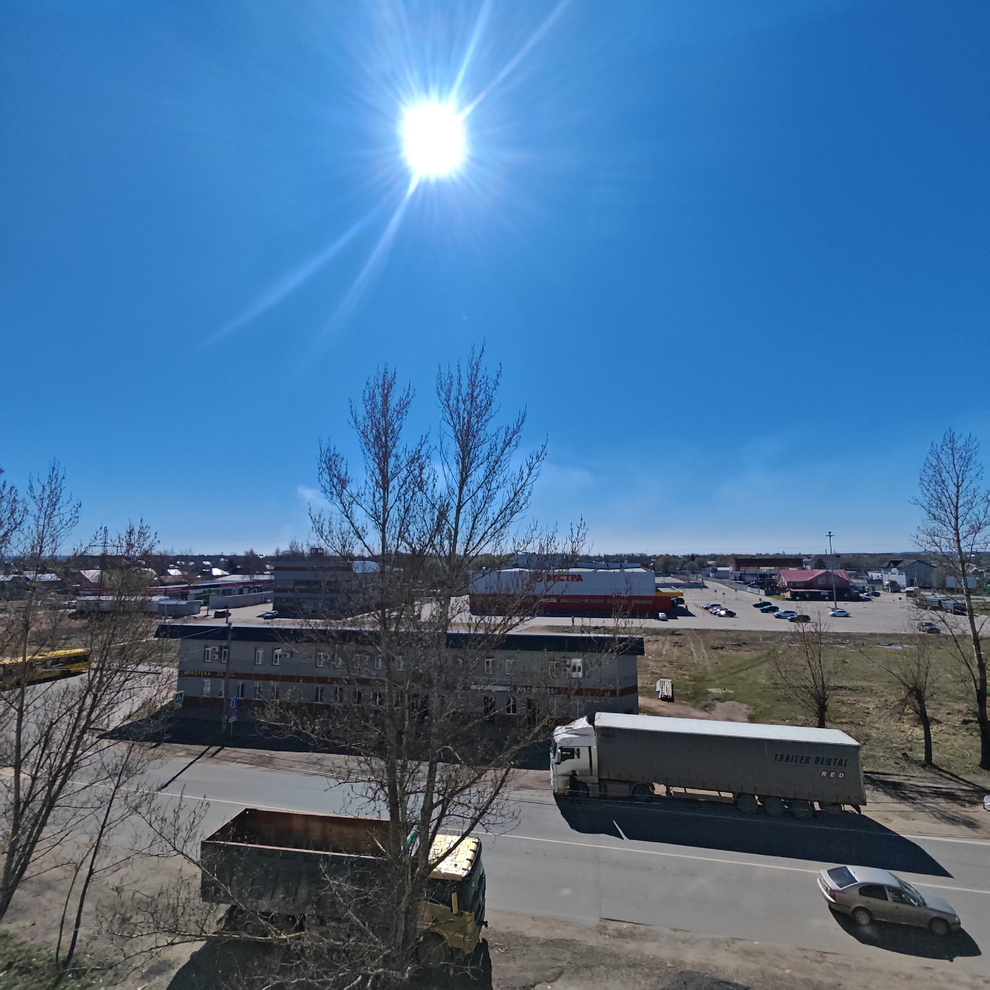 The image depicts an urban scene under a bright, sunny sky. The sun is high in the sky, creating significant glare. The scene is dominated by a road with vehicular traffic, including two trucks and a car. One truck is a dump truck, the other is a trailer truck. A building complex is visible in the background, with a large store and some smaller businesses and buildings. A large lot is on the far side of the commercial properties. Bare trees are scattered throughout the image, suggesting it is early spring or late autumn. The location is Rostov, Russia.