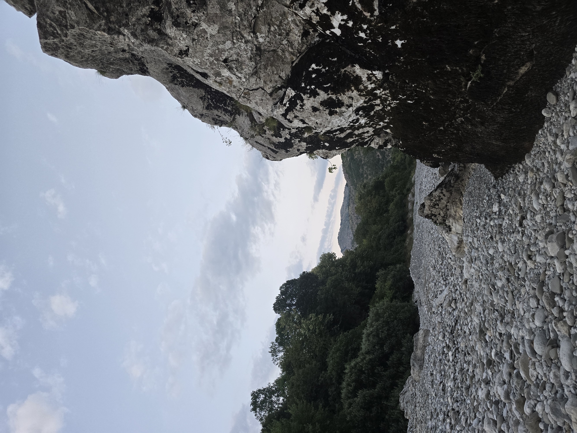 This image depicts a rocky, dry riverbed with a large rock formation dominating the right foreground. The riverbed is covered in numerous rounded grey stones of varying sizes, stretching into the distance. To the left, a dense treeline of lush green foliage lines the bank, with further hills visible beyond. The sky above is a pale blue, with scattered, soft grey clouds. The overall impression is a natural, undeveloped landscape. The location is identified as Bioče, Montenegro. There are no people or animals visible in the image.