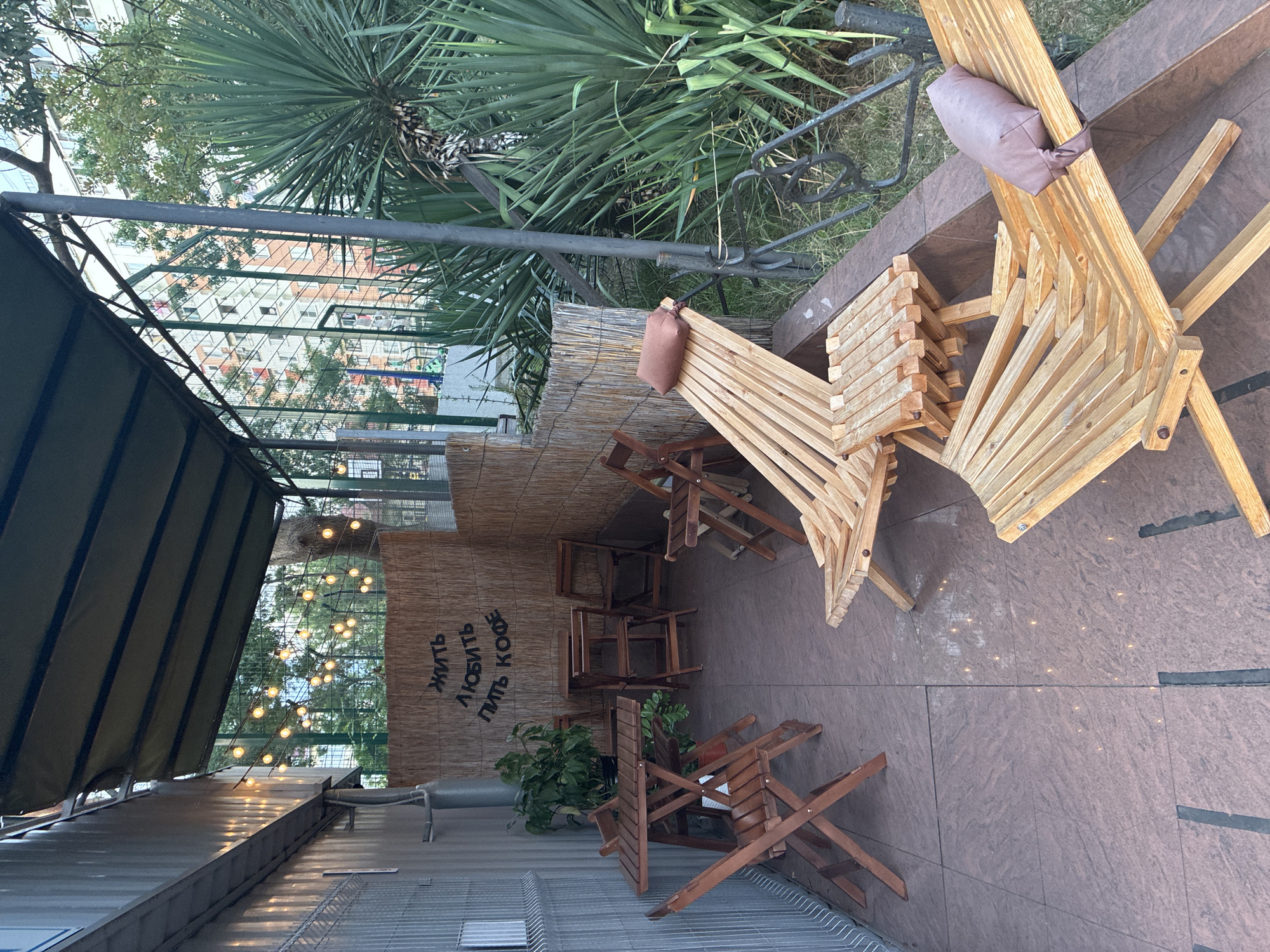 The image depicts an outdoor seating area, likely a cafe patio, situated in Sochi, Russia. The scene is framed by a corrugated metal wall on the left, a thatched bamboo-like screen in the middle, and lush green foliage, including a large yucca plant, on the right. String lights are visible, illuminating the covered area.
In the foreground, several wooden deck chairs and a small wooden table are arranged on a tiled floor. One of the deck chairs has a mauve pillow resting on it. Further back, there is a set of wooden folding tables and chairs. A plant in a pot sits near the tables.
Above the seating area, a dark green awning provides shade. In the background, a tall, light-colored building with many windows and balconies is visible, along with a green chain-link fence and trees. The overall impression is of a casual, relaxed outdoor space.