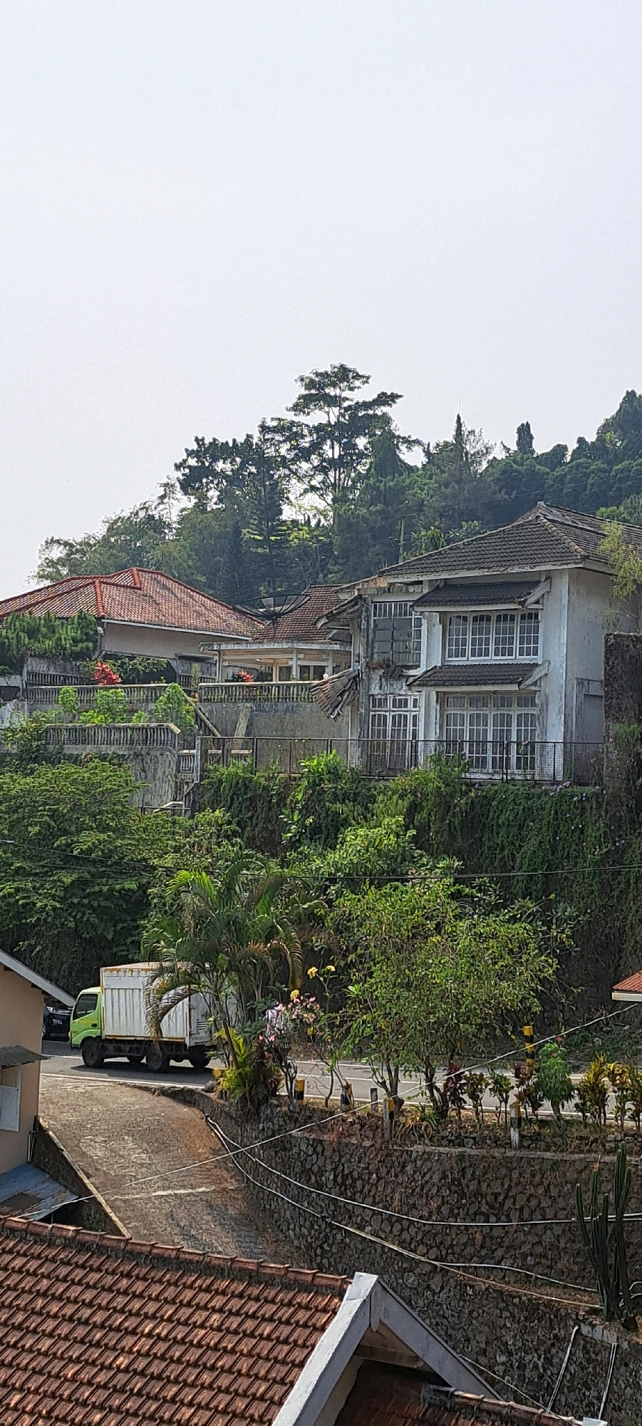 The image depicts a hillside landscape in the city of Prigen, Indonesia, characterized by several residential structures nestled amongst dense foliage. In the foreground, a section of a terracotta-tiled roof is visible, suggesting a residential area. A paved road winds uphill, with a light green truck in motion. Further up the hill, there are multiple buildings. One building features a red tile roof, while another, larger white building exhibits multiple stories with numerous windows and a weathered appearance. Extensive vegetation, including trees and various shrubs, covers the hillside, creating a lush, green environment. A stone retaining wall runs along the side of the road, supporting the elevated terrain and gardens. The sky is overcast, contributing to a diffused natural light.