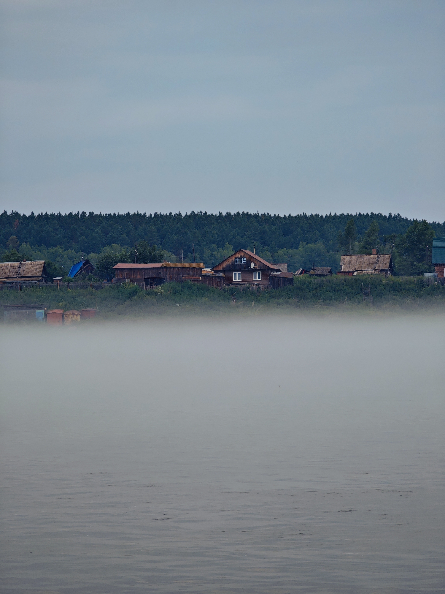 The image depicts a foggy morning scene along the banks of a body of water, likely a river, in the village of Atamanovo, Russia. In the foreground, the water's surface is visible, characterized by gentle ripples and a muted gray color. A thick layer of fog obscures the lower portion of the scene, creating a hazy transition between the water and the land.
Beyond the fog, on the elevated bank, a collection of rustic wooden houses and other structures are visible. These buildings appear to be older, with weathered wooden facades and a mix of pitched and flat roofs. Some of the houses have windows, and one house on the left has a distinct red-tiled roof. There are also what appear to be storage containers or small structures in a rust color located near the water's edge, partially shrouded by vegetation.
Behind the village, a dense forest of dark green coniferous trees forms a backdrop, stretching across the horizon. The sky above is overcast and a pale gray, contributing to the subdued and slightly somber atmosphere of the scene. The overall impression is one of a quiet, rural settlement on a misty morning.