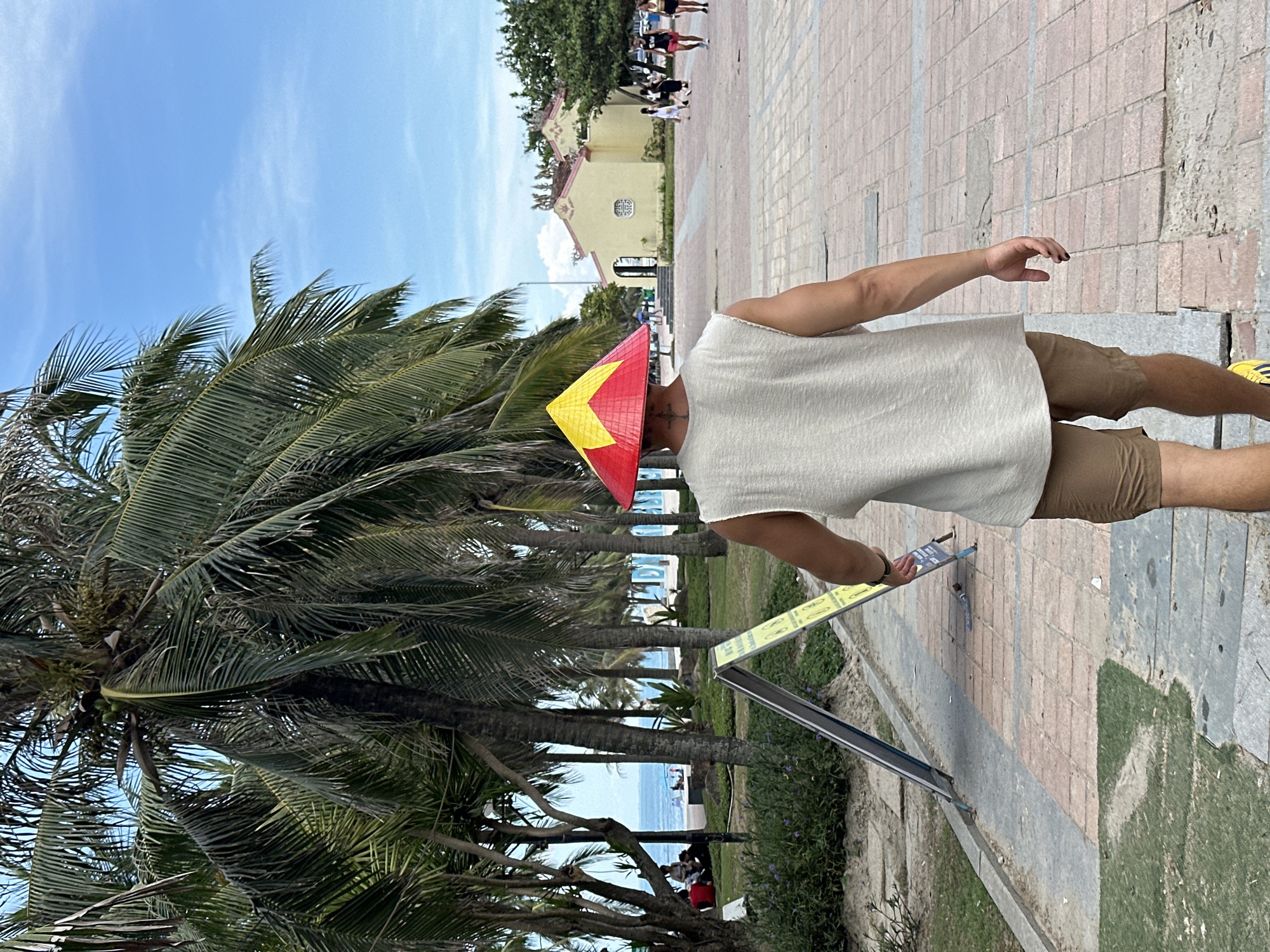 A person wearing a conical hat with red and yellow stripes is standing on a paved walkway lined with palm trees. The person is wearing a light-colored, sleeveless shirt and brown shorts. They are holding a small object in their right hand, possibly a phone. In the background, a building with a yellow facade and ornate roofline is visible, along with other people walking on the path. Further back, the ocean can be seen, with a few people relaxing on the beach. The sky is blue with wispy clouds. The scene suggests a sunny day in a tropical location, likely a tourist area by the beach. The orientation of the image is landscape, but the content is rotated.