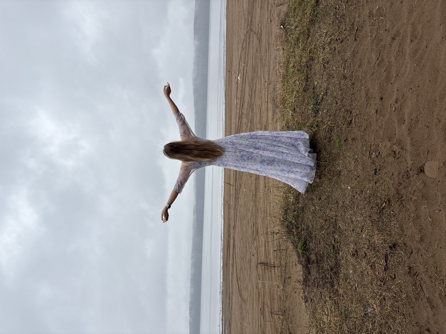 The image depicts an outdoor scene featuring a young woman standing on the shore of a large body of water under a cloudy sky. The woman, seen from behind, is positioned slightly off-center to the right. She has long, straight blonde or light brown hair that extends down her back. She is wearing a flowing, light-colored dress, possibly with a floral or subtle pattern, and sheer sleeves. A dark wristwatch is visible on her left wrist. Her arms are outstretched sideways and slightly upwards, a pose that conveys a sense of freedom, openness, or embrace of the environment.
The foreground consists of a sandy or dirt bank with patches of dry, sparse grass and weeds. Some faint marks, possibly tire tracks or footpaths, are visible on the sandy surface. Beyond the immediate shore, a wide and calm body of water stretches across the middle ground, reflecting the soft, diffused light of the sky. In the distance, across the water, a low, dark line suggests a distant landmass or horizon.
The sky, which occupies the upper portion of the frame, is entirely overcast and grey, filled with soft clouds, indicating a cloudy day. The overall lighting is subdued and even. The scene evokes a tranquil and expansive mood. There is no visible text in the image. Given the location context of Chaikovsky, Russia, the body of water is likely a large river or reservoir, such as the Kama River or Votkinsk Reservoir.