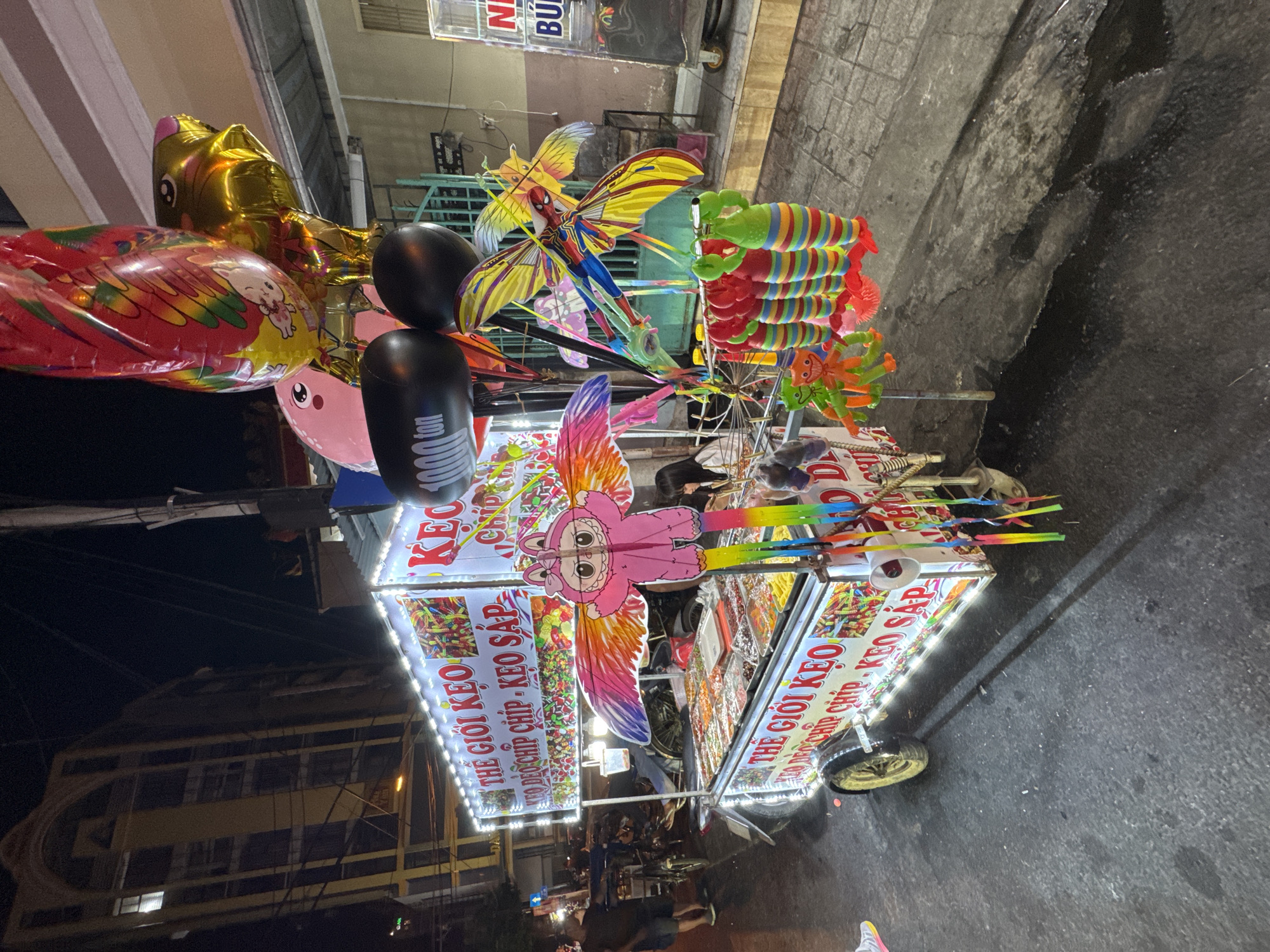 An outdoor scene at night shows a candy cart in Nha Trang, Vietnam. The cart is illuminated by bright white LED lights and displays a variety of colorful candies in clear containers. The cart is decorated with large, brightly colored banners that read "THE GIOI KEO" and "KEO CHIP CHIP - KEO SAP," indicating it sells candy. Several large, colorful balloons, including animal-shaped ones, are attached to the top of the cart. Hanging from the cart are various toys, including kites shaped like butterflies and a figure resembling Spider-Man.
A person wearing a dark shirt and shorts is visible on the left side of the frame, standing near a bicycle. Another person, a young woman with dark hair, is seated behind the cart on the right side, partially obscured by the display. The cart is on a paved street, with a dark, wet asphalt surface in the foreground, suggesting recent rain or cleaning. In the background, a multi-story building with arched windows is visible, its facade illuminated. The overall atmosphere suggests a lively night market or street vendor setup.
