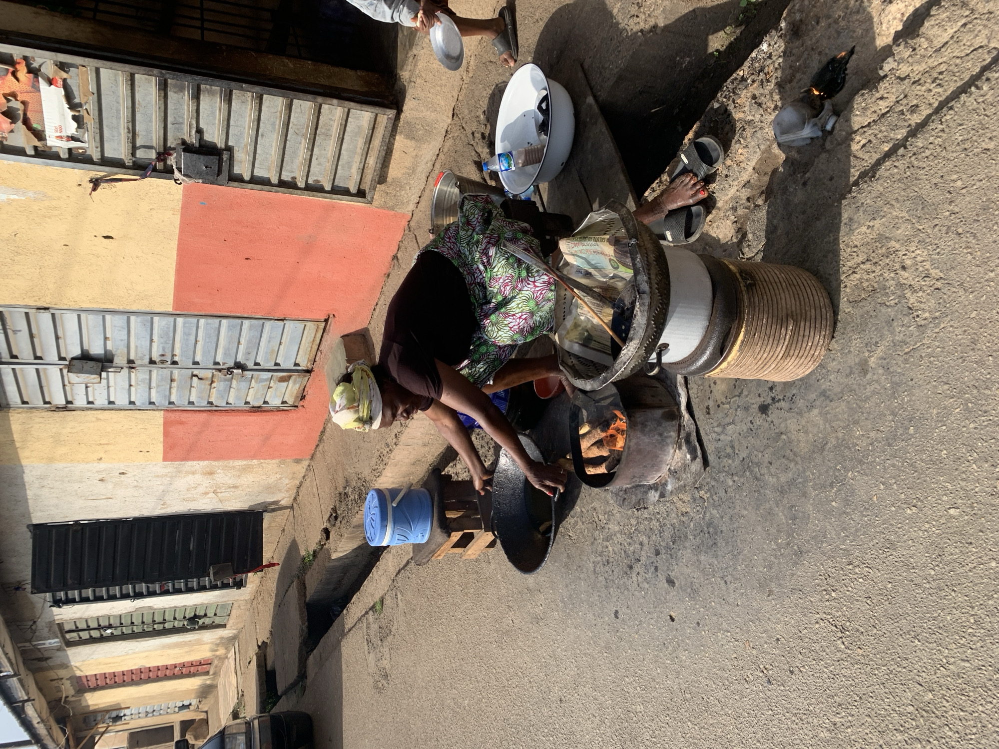 An elderly woman in Jos, Nigeria, cooks outdoors on a makeshift stove, likely in a street or alley. She wears a headscarf, a dark-colored shirt, and a colorful patterned skirt. She is bending over a large, black cooking pot, positioned over a fire fueled by wood in a crude stove. Her hands are visible, manipulating something within the pot. To her left, there is a blue plastic container and a stack of wooden blocks. Behind her is a woven basket overflowing with what appears to be money, possibly Nigerian Naira notes. The background includes buildings with metal doors, painted in alternating sections of yellow and red. In the upper part of the frame, other people are partially visible. The ground is paved, with indications of dirt and debris. A second fire is burning outside the stove.